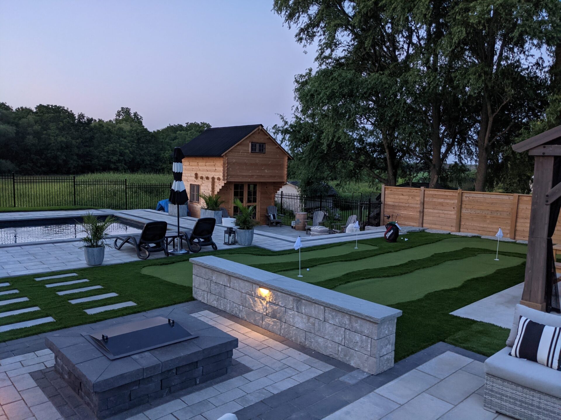 An outdoor space at dusk featuring a rectangular pool, artificial putting green, a fire pit, seating areas, and a wooden shed. A person is visible.