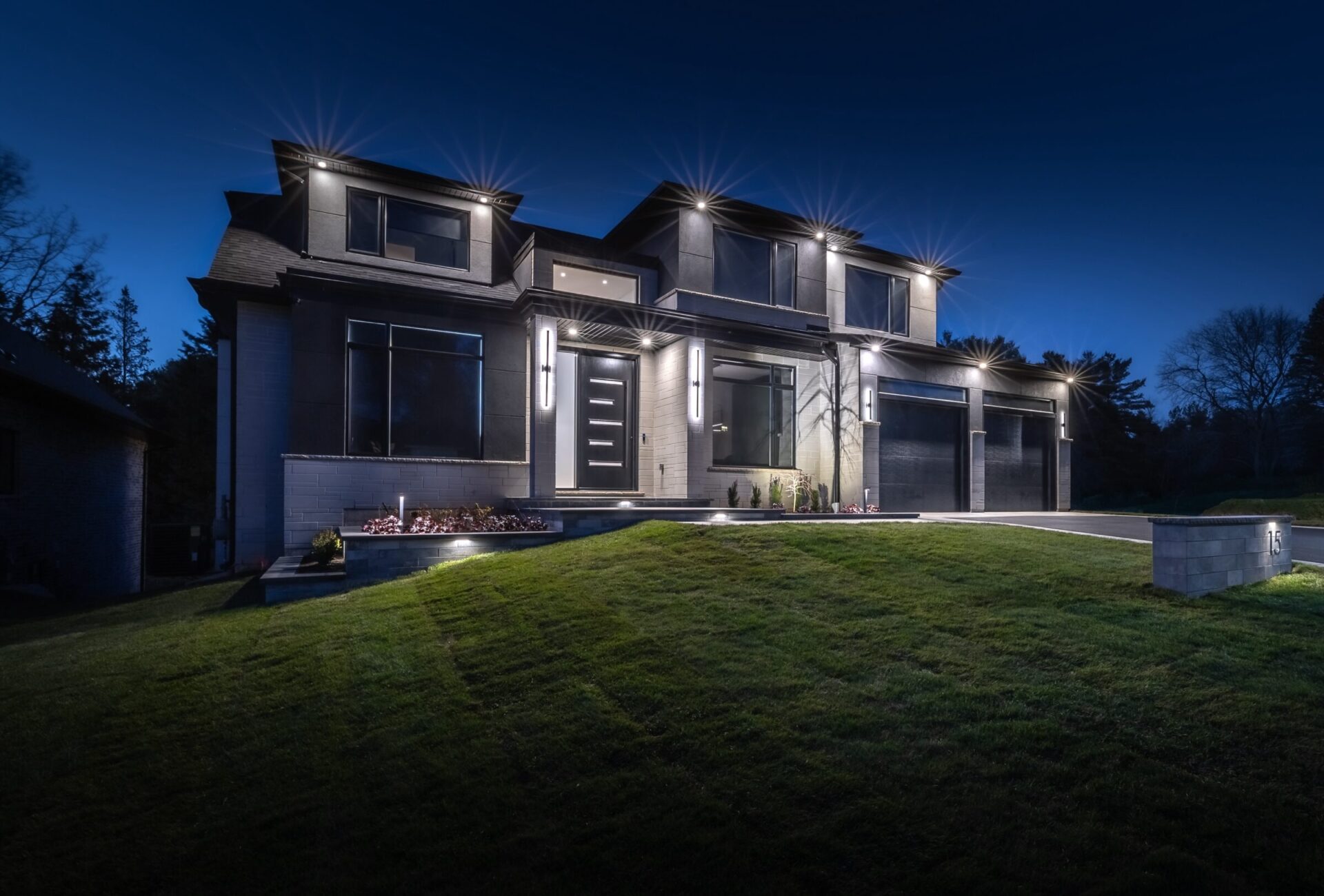 A modern two-story house at dusk with exterior lights on, showcasing large windows, a flat roof, landscaped garden, and a well-manicured lawn.