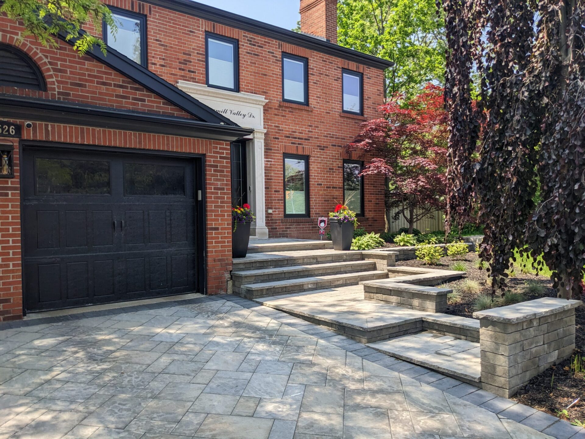 This is an image of a brick house with a black garage door, stone paved driveway, and steps leading to the entrance surrounded by landscaping.