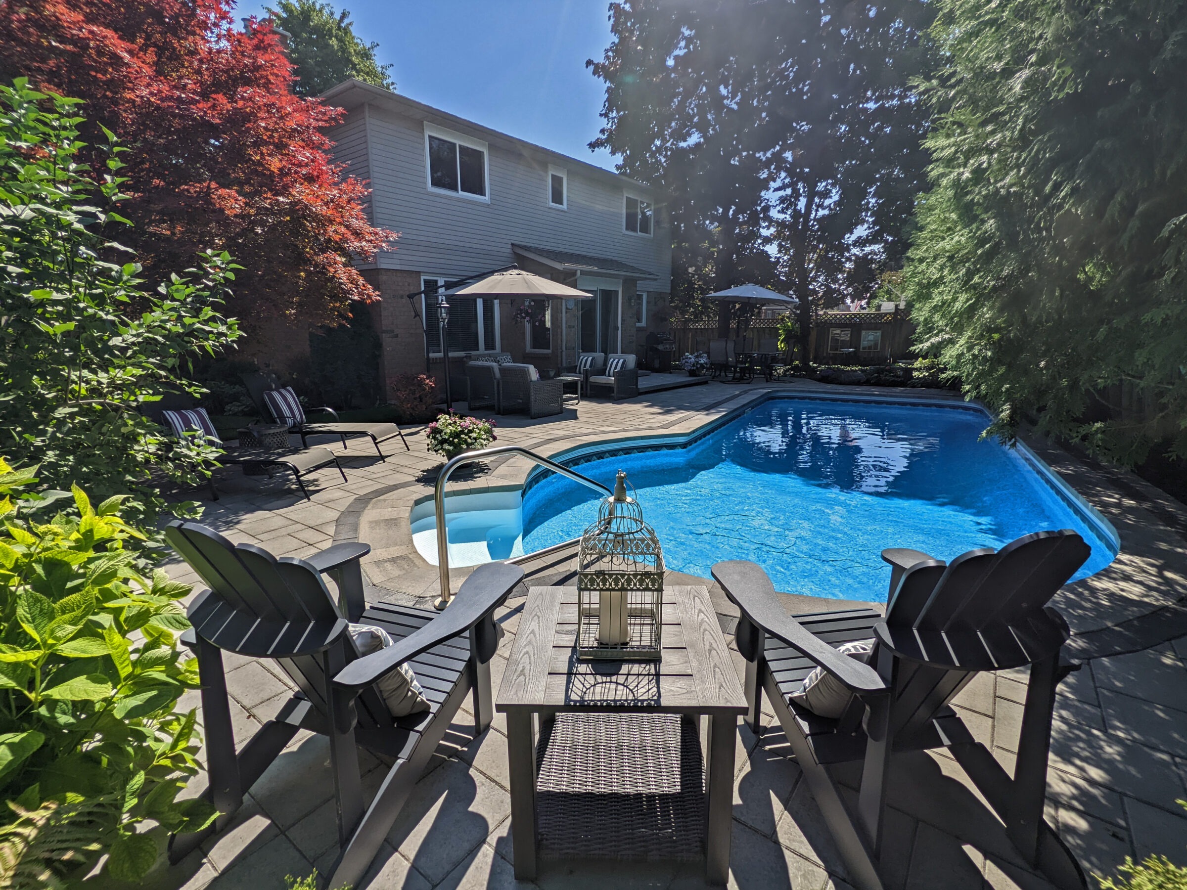 A sunny backyard with a blue swimming pool, patio furniture, lush greenery, a two-story house, and a clear blue sky.