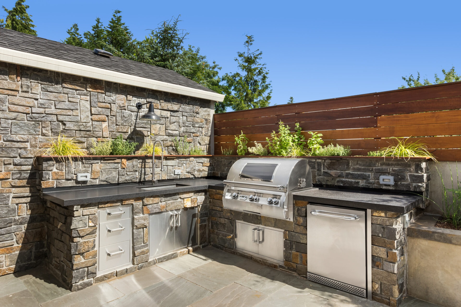 Outdoor kitchen with stone facade, stainless steel appliances, and a grill; set against a backdrop of trees and a wooden fence, under a clear sky.