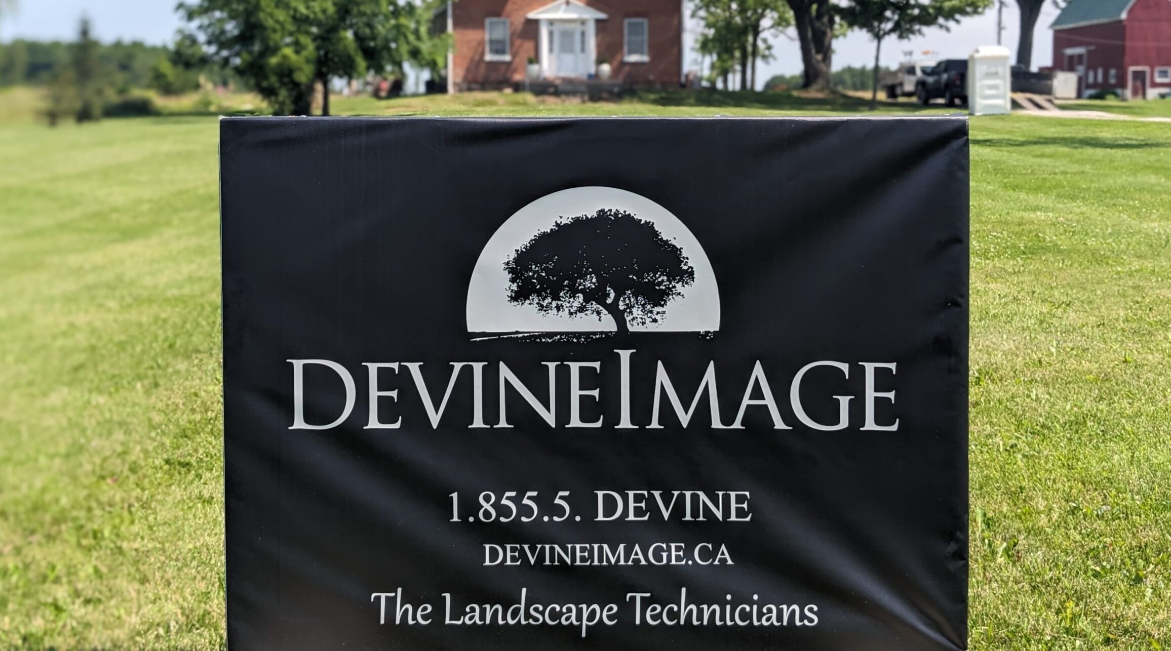 A black sign with white text reads "DEVINE IMAGE," displaying a tree logo, contact information, and the tagline "The Landscape Technicians," set against a grassy background.