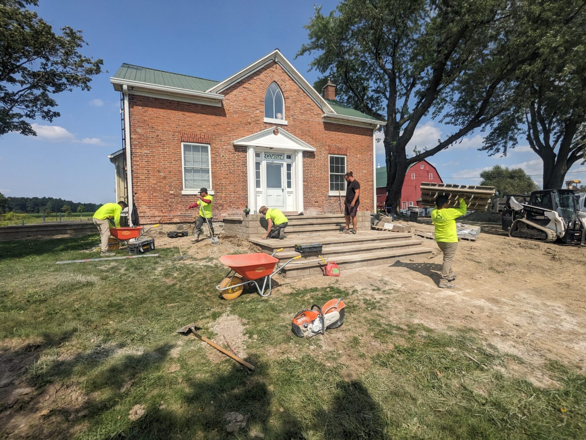 A group of people in safety vests work on a wooden deck outside a brick building with a green metal roof, near construction equipment and tools.