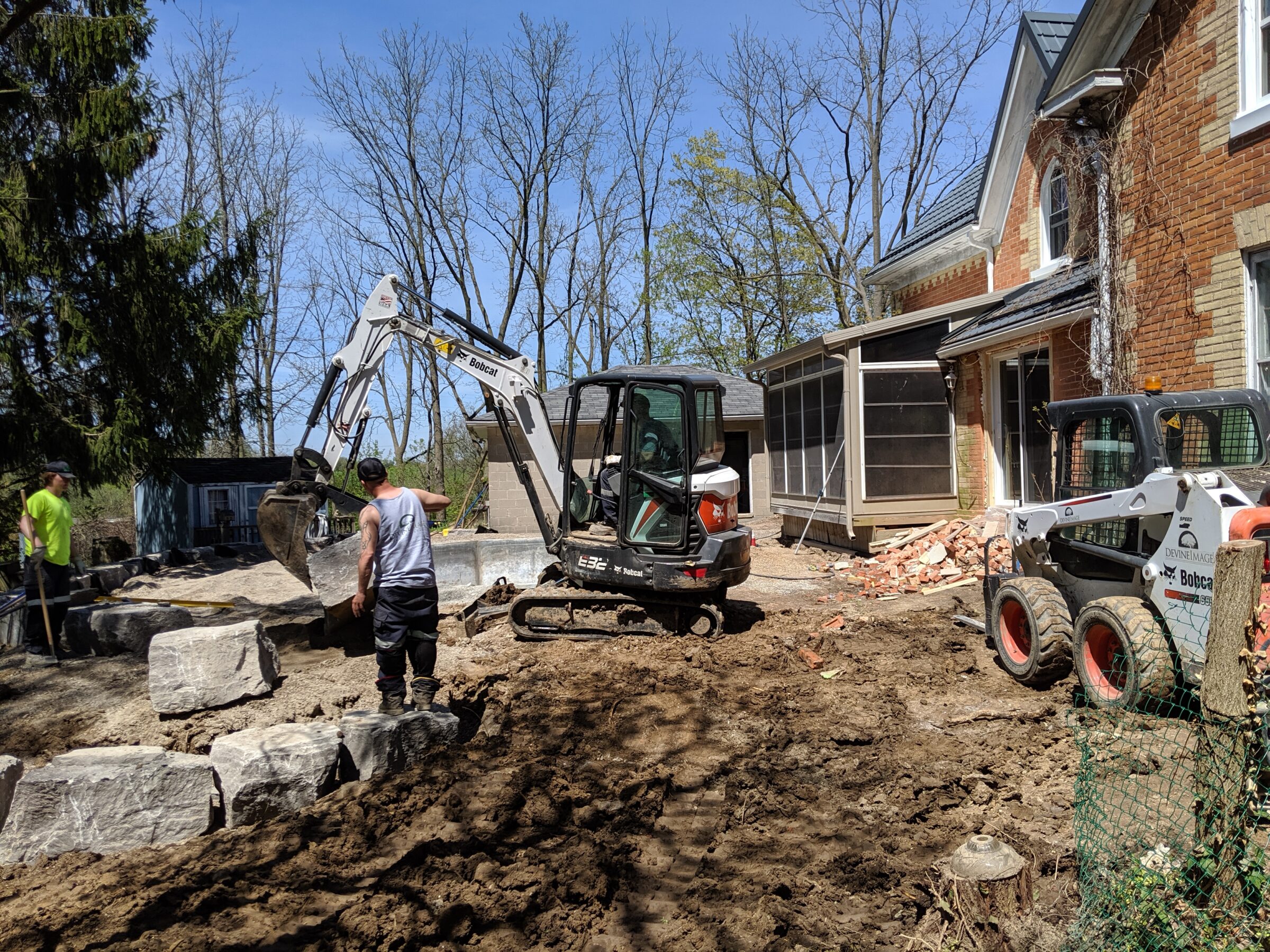 In a residential construction site, two people work near heavy machinery, handling large stones amidst debris, under a clear sky by a brick house.