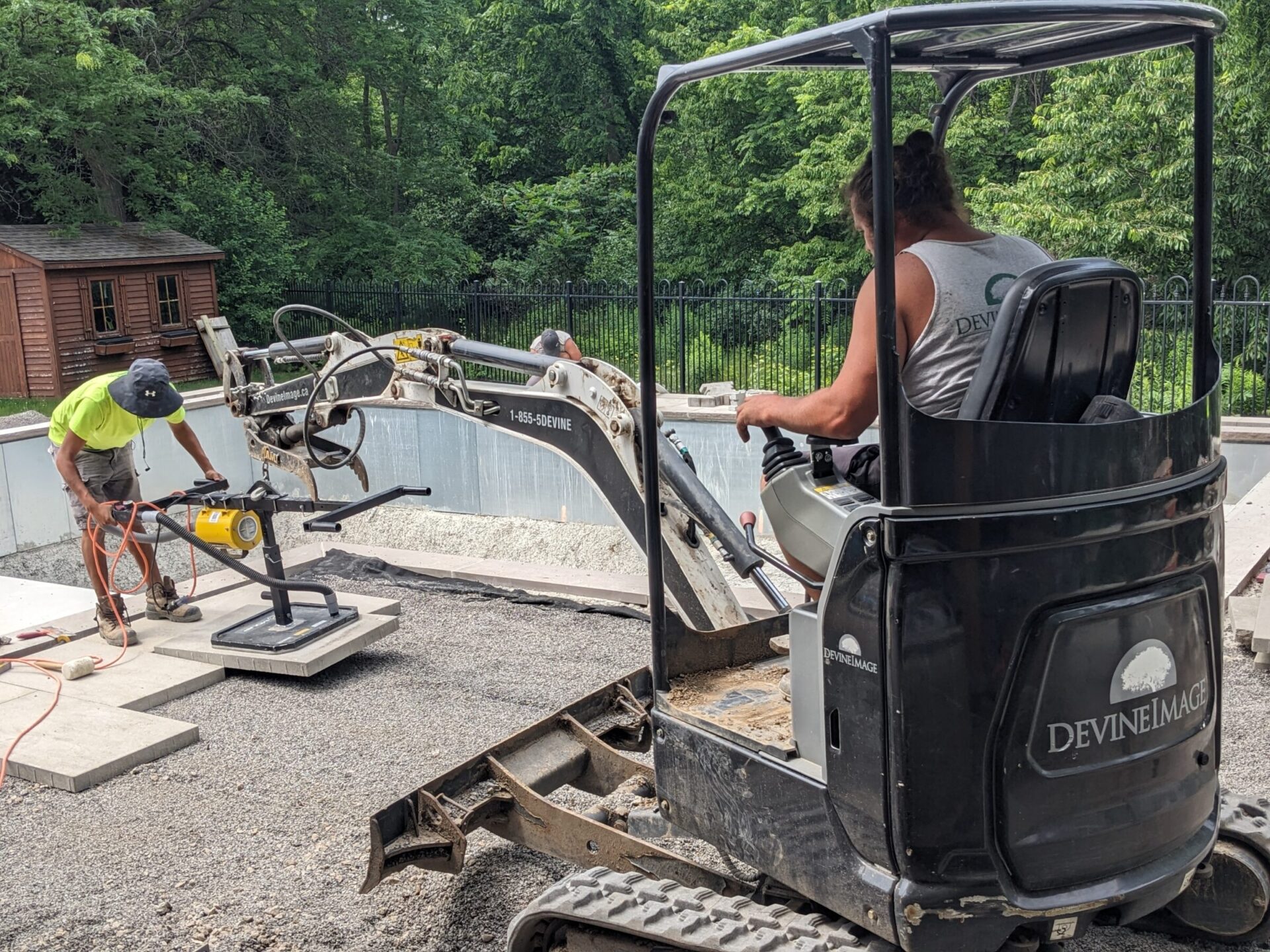 Two people are working at a construction site near a pool; one operates machinery while the other cuts material, with trees and a shed in the background.