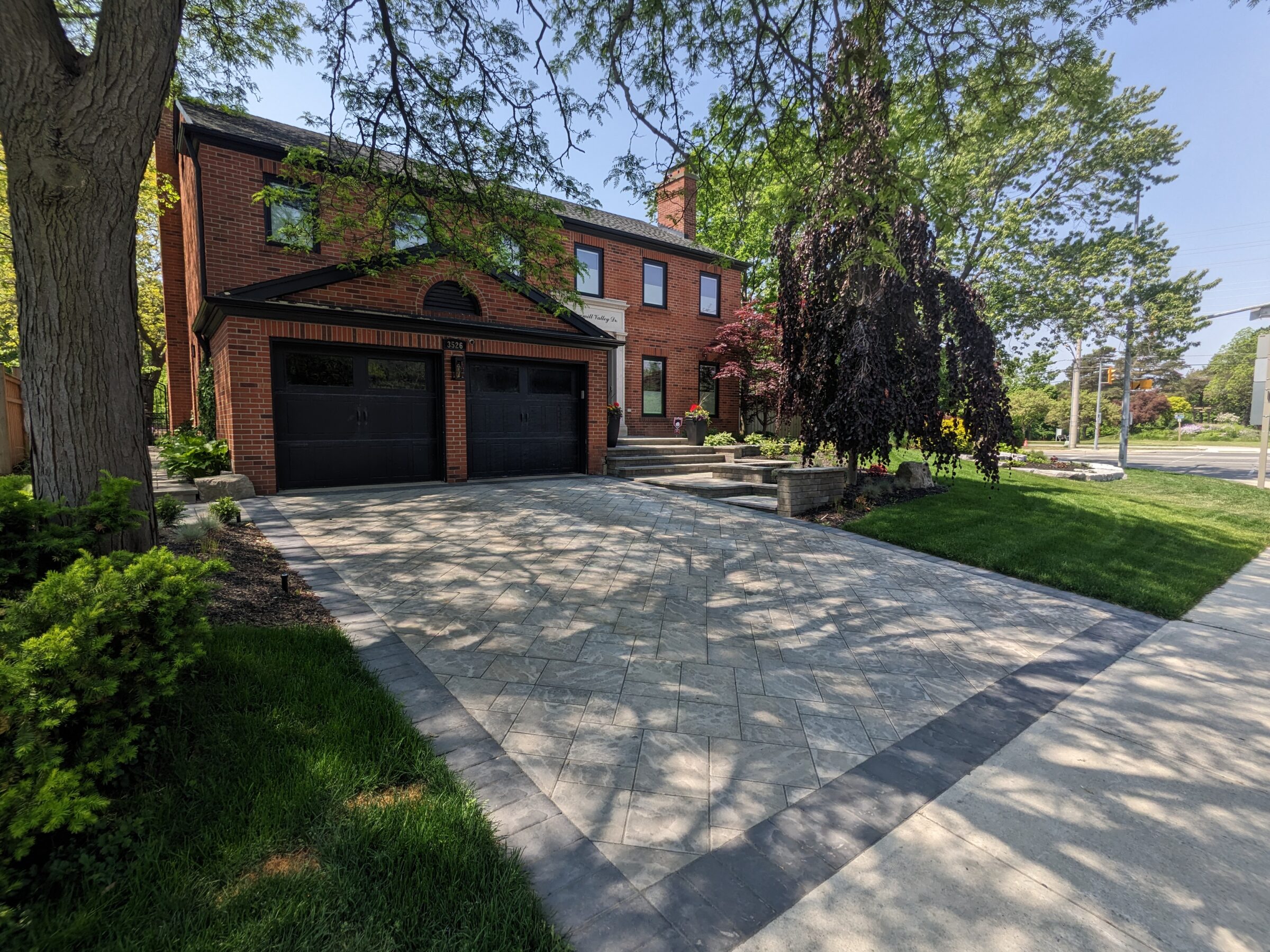 A brick house with a double garage door, patterned driveway, and well-landscaped front yard under a clear sky, surrounded by trees and greenery.