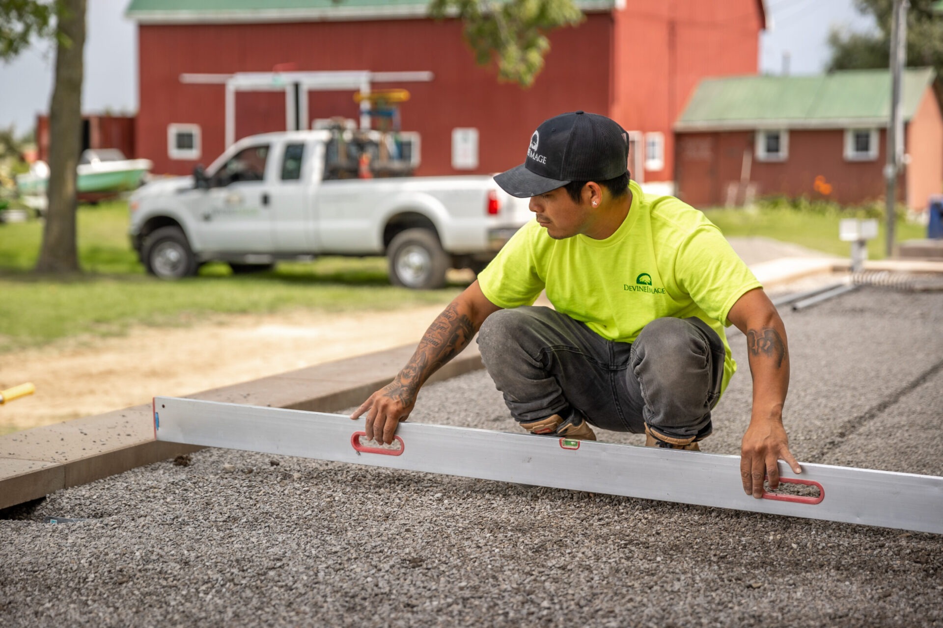 A person is kneeling on gravel, using a long level on a concrete structure, focused on their work, with a truck and red barn in the background.