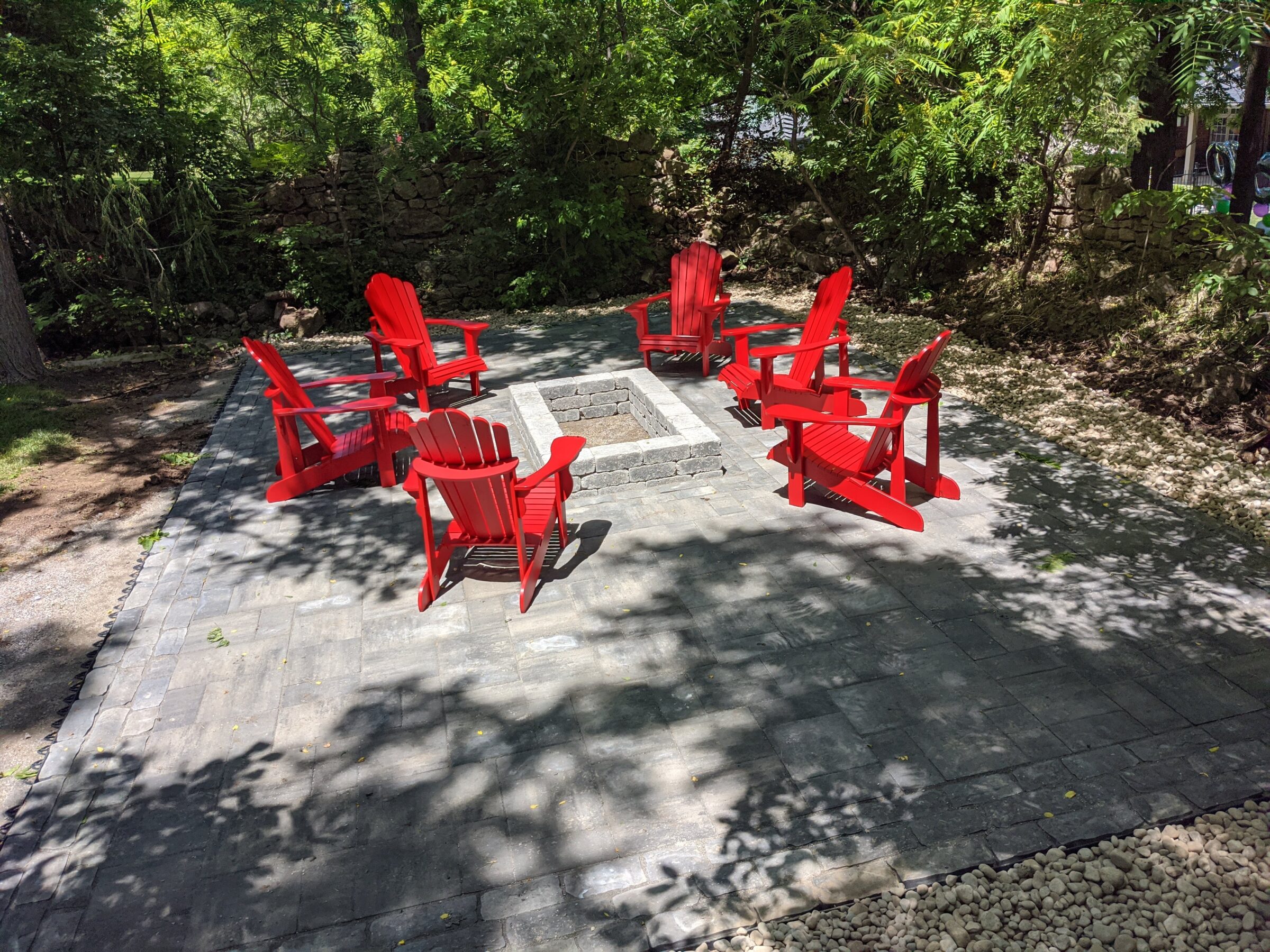 A serene outdoor setting with four red Adirondack chairs arranged around a square fire pit, on a patio with surrounding greenery and dappled sunlight.