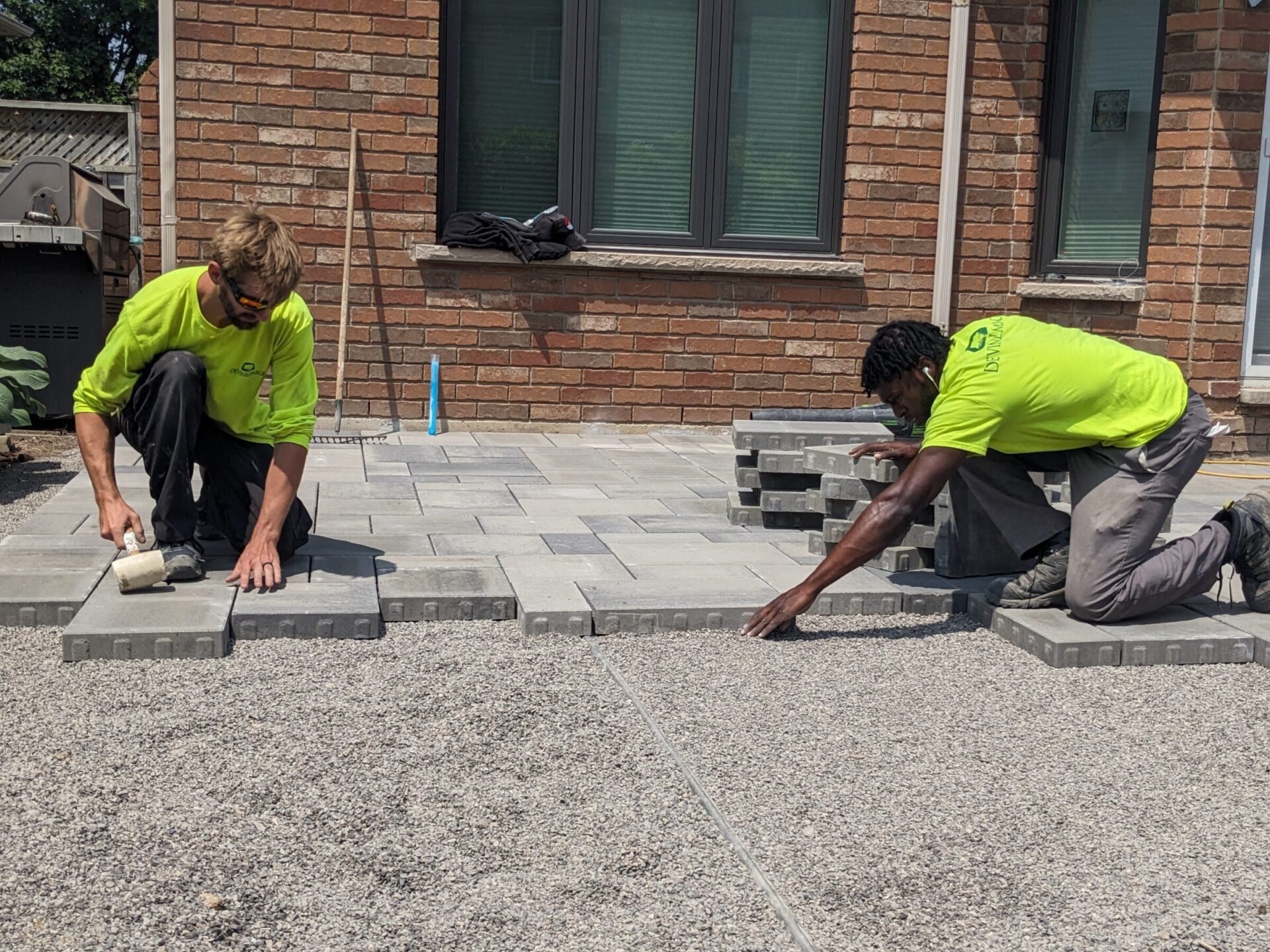 Two people in bright green shirts are installing grey paving stones on a driveway near a brick house on a sunny day.