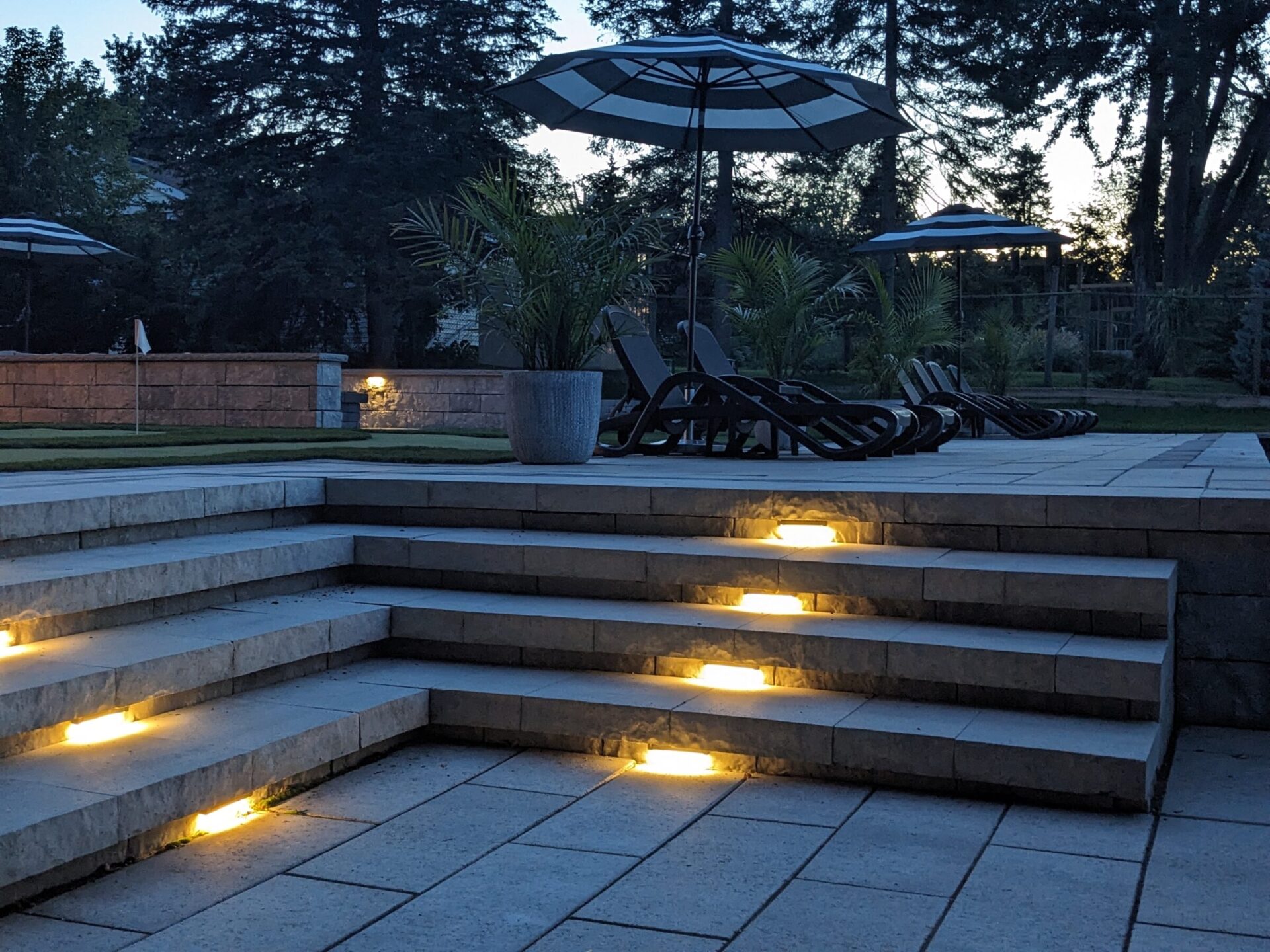 An outdoor patio at dusk with lit step lights, sun loungers, umbrellas, trees, and a well-manicured lawn in a tranquil setting.
