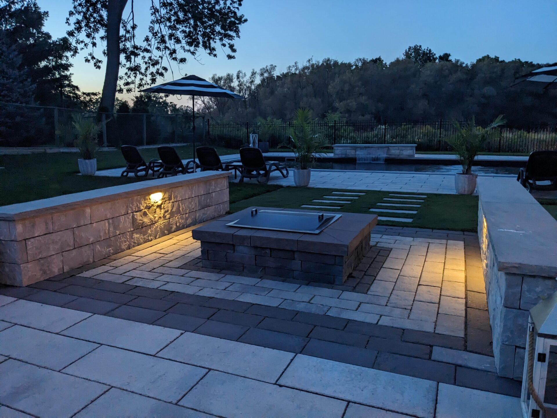 Twilight setting over a landscaped backyard with a paver patio, sun loungers, an umbrella, an outdoor kitchen, and soft built-in lighting. Trees line the background.