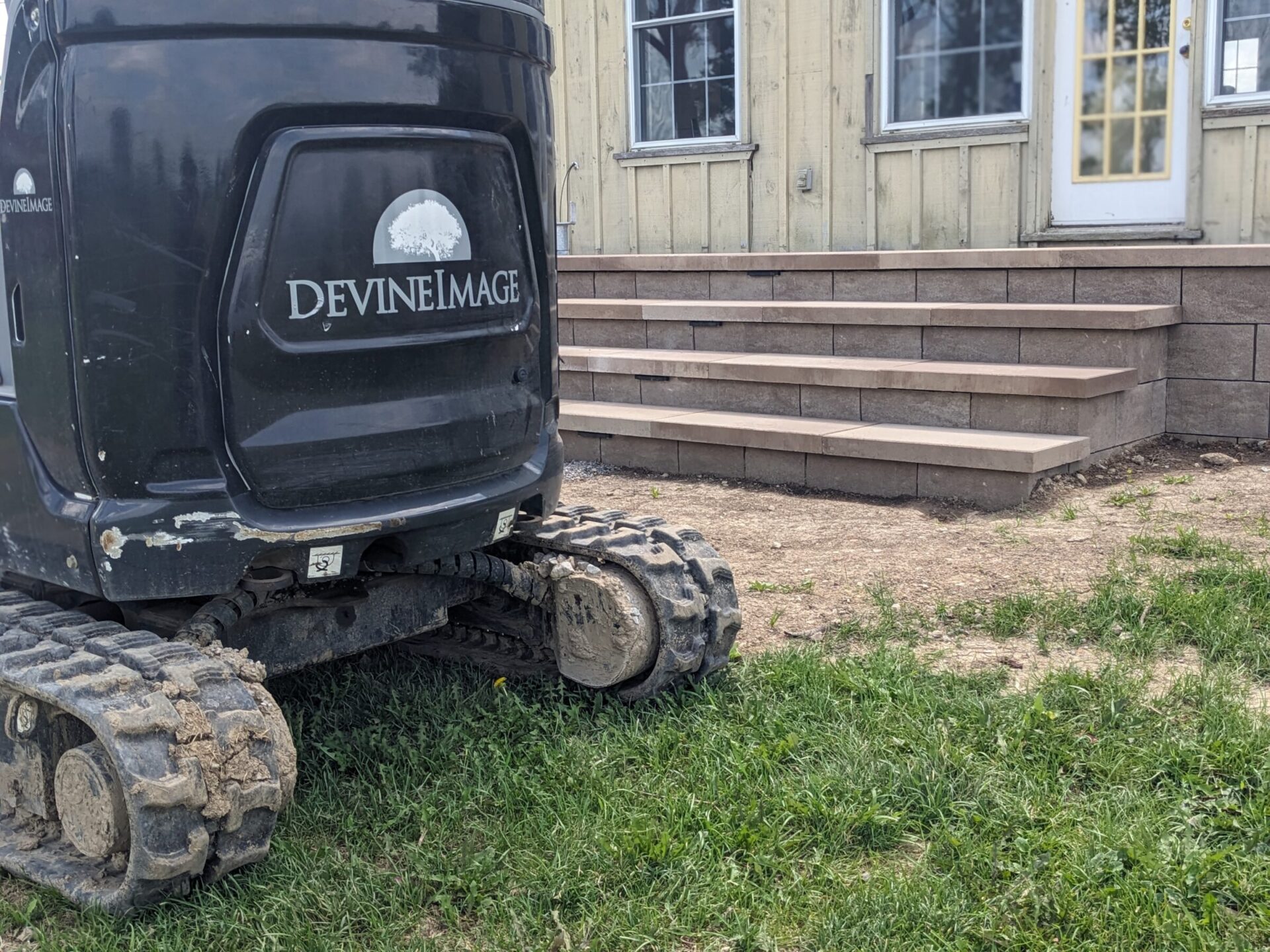 The image shows a compact track loader with the logo "DEVINE IMAGE" on its side, parked next to a building with a newly built stone staircase.