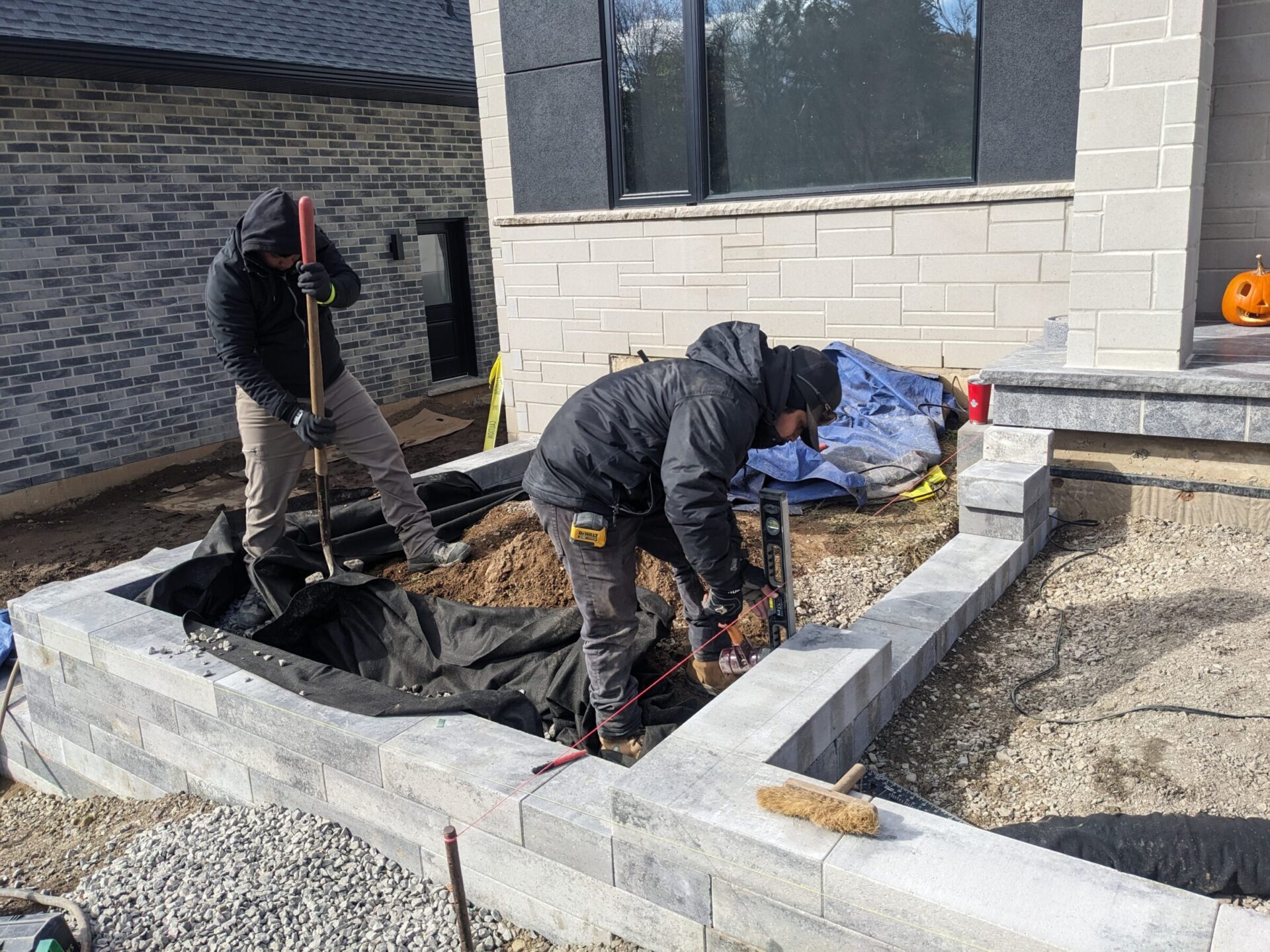 Two people are working on landscaping outdoors, shoveling dirt near a stone retaining wall, under clear skies by a building with brick walls.