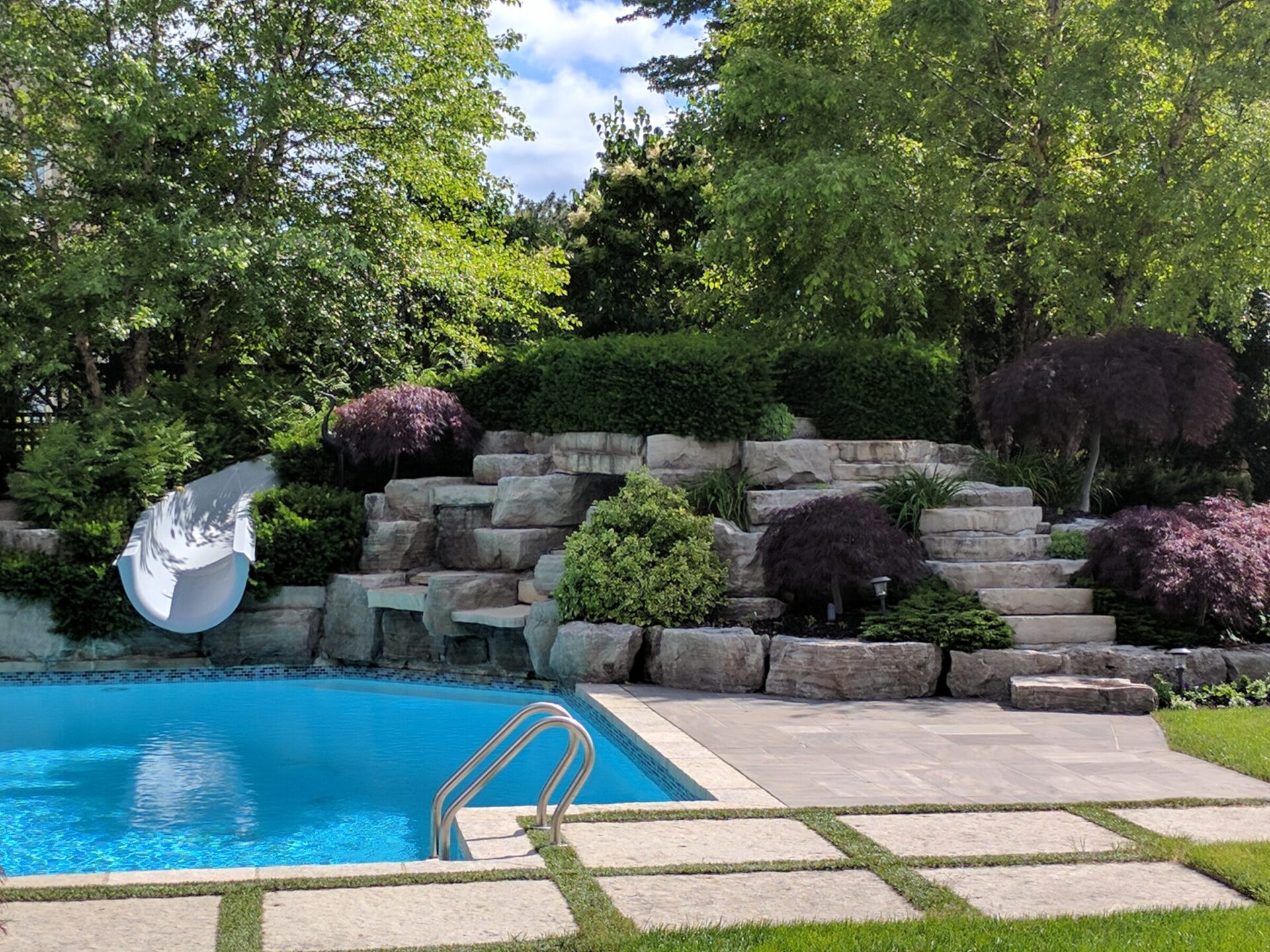 A backyard with a blue swimming pool, stone steps, a white slide, landscaped with various plants and trees under a sunny blue sky.