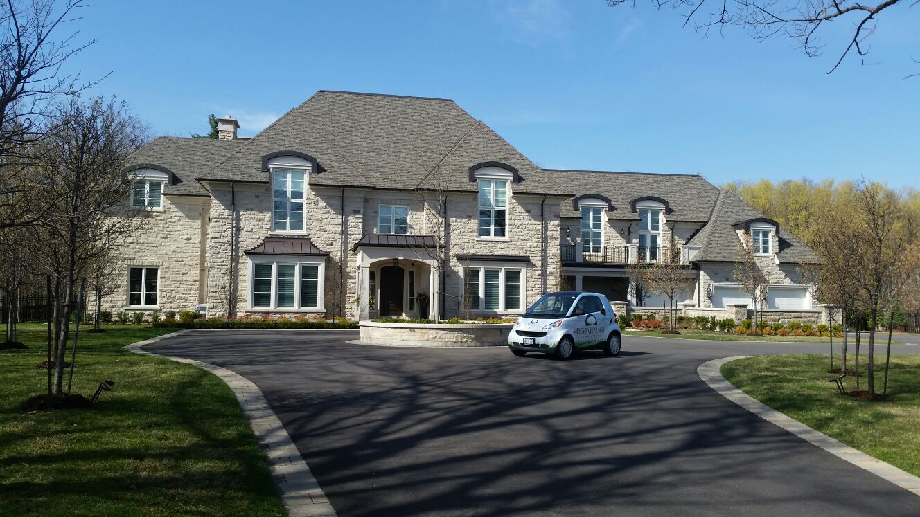 This image shows a large, two-story estate with stone walls and a shingled roof on a sunny day, featuring a landscaped yard and a small car.