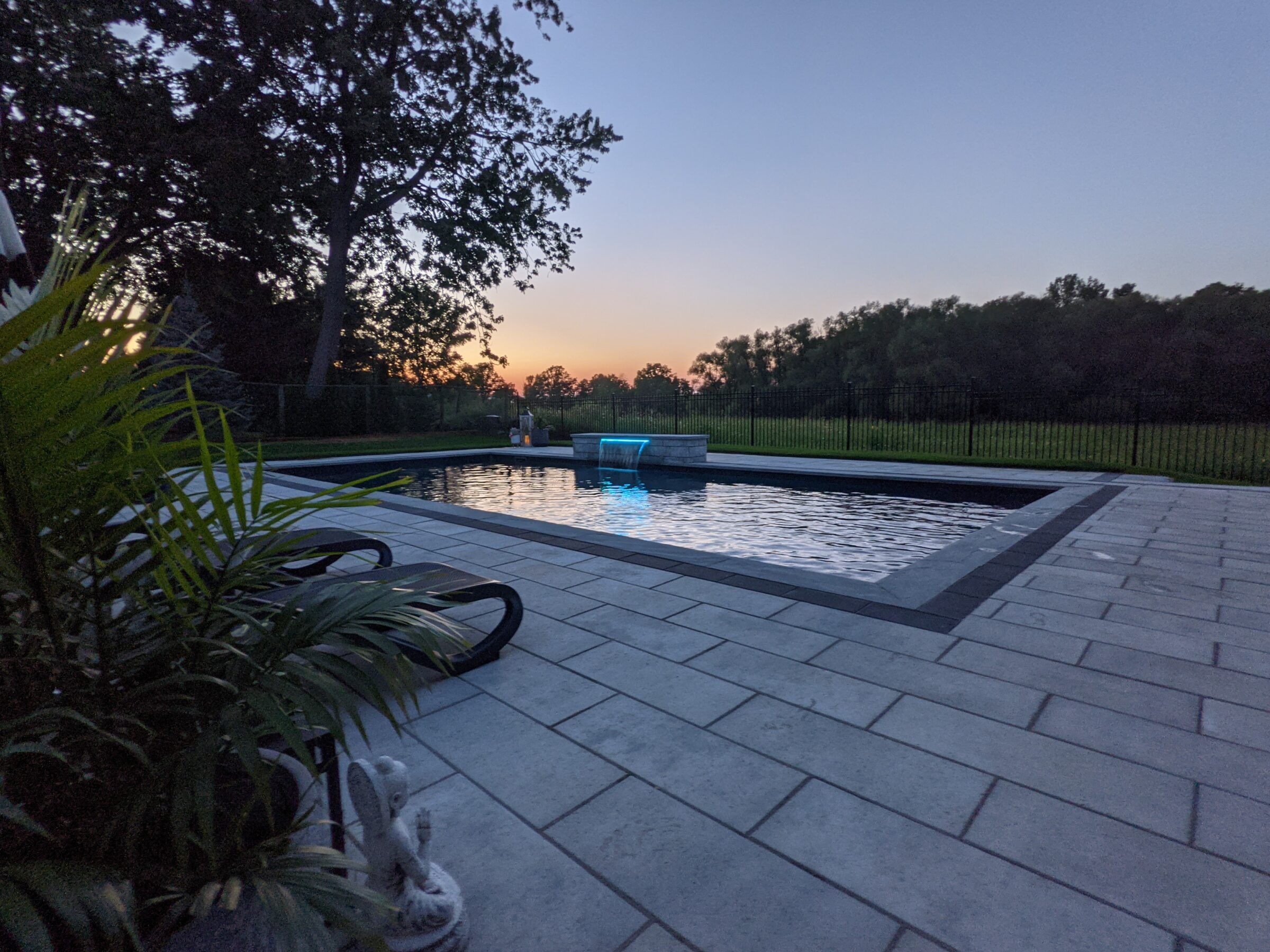 An outdoor swimming pool at dusk with waters reflecting the sky's colors, surrounded by paving stones and flanked by greenery and a fence.