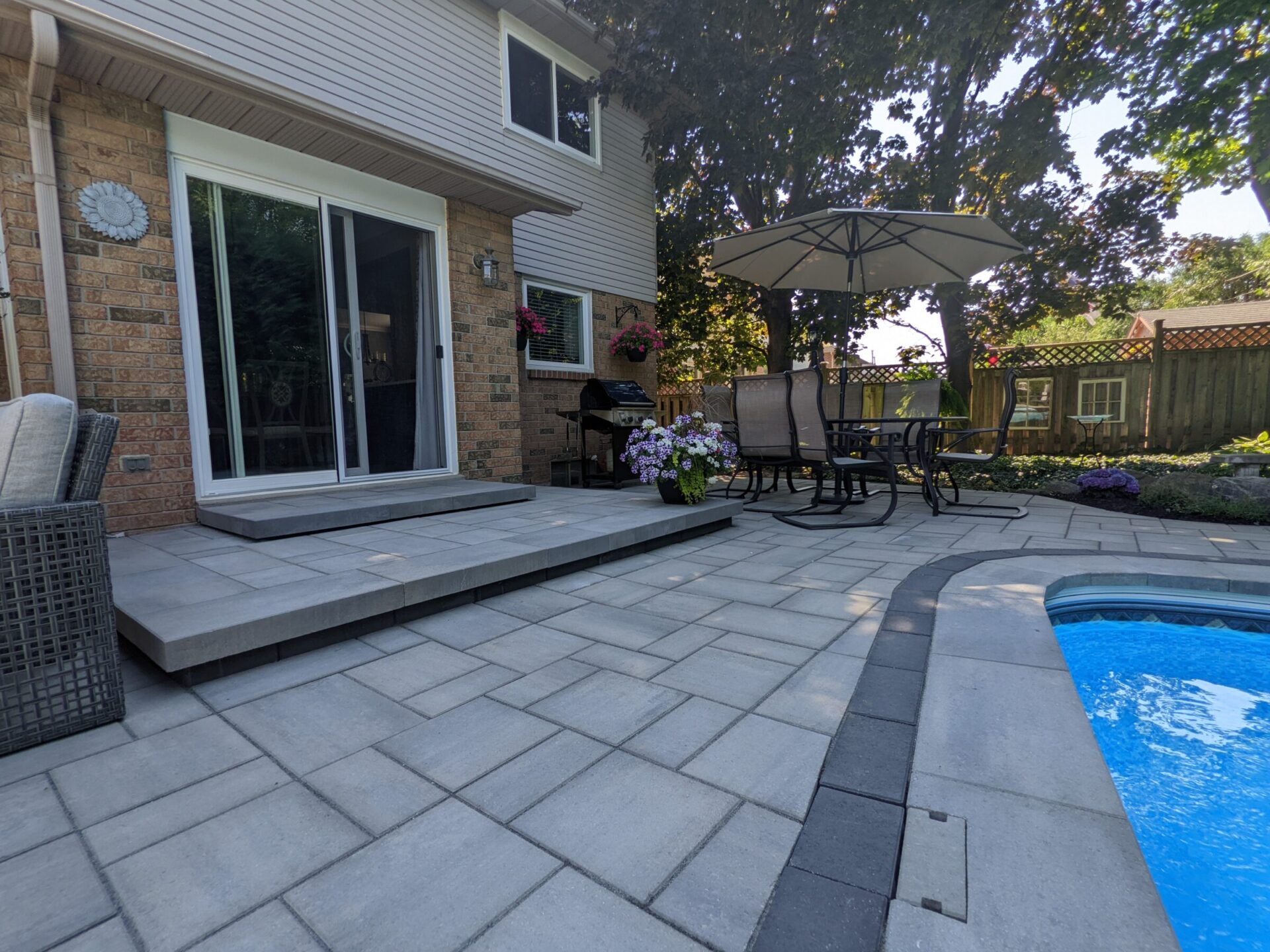 This image shows a backyard patio with paving stones, a dining set under an umbrella, potted plants, a BBQ grill, part of a swimming pool, and trees.