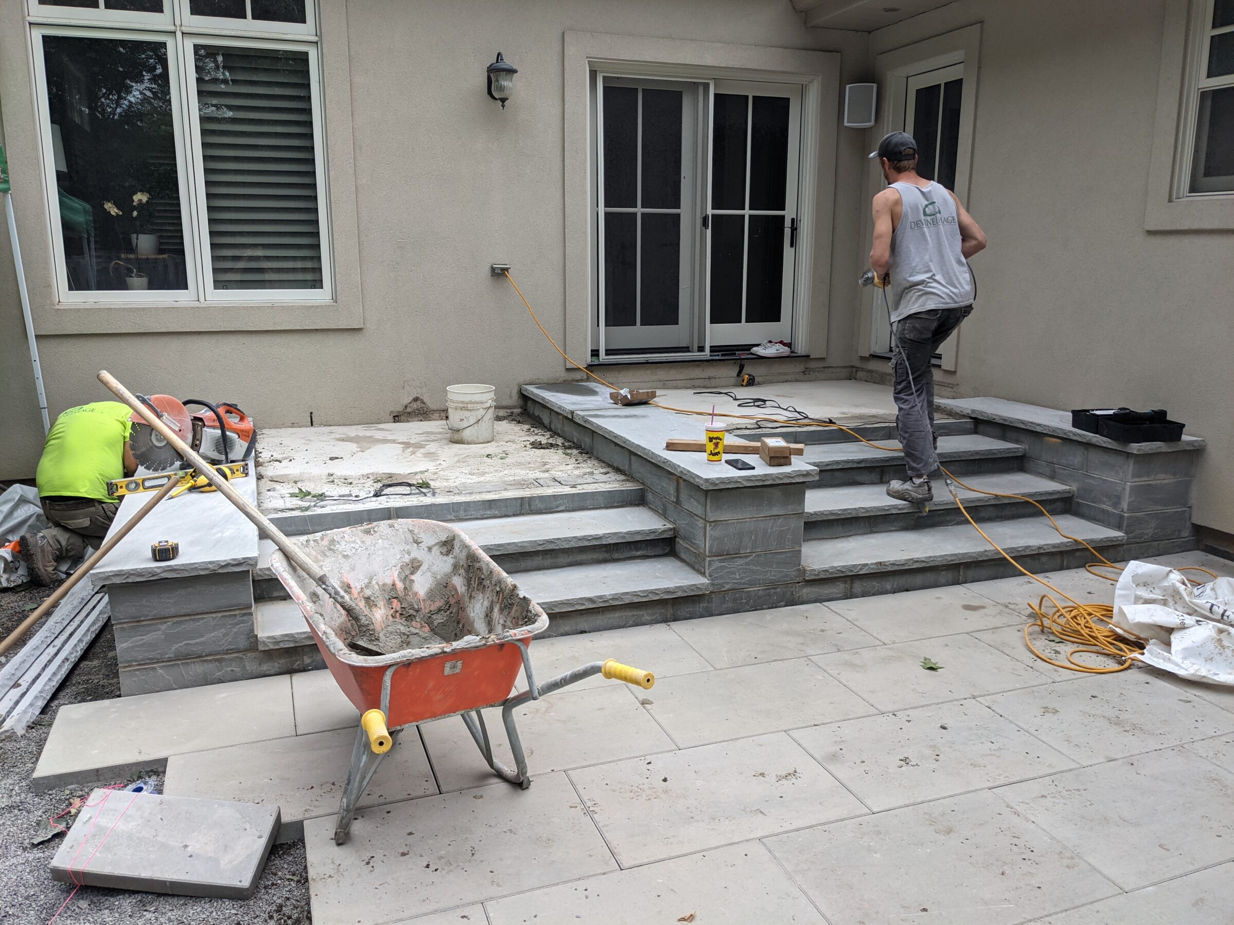 This image shows two people working on a patio construction with stone steps. There's a wheelbarrow, tools, and building materials scattered around the site.