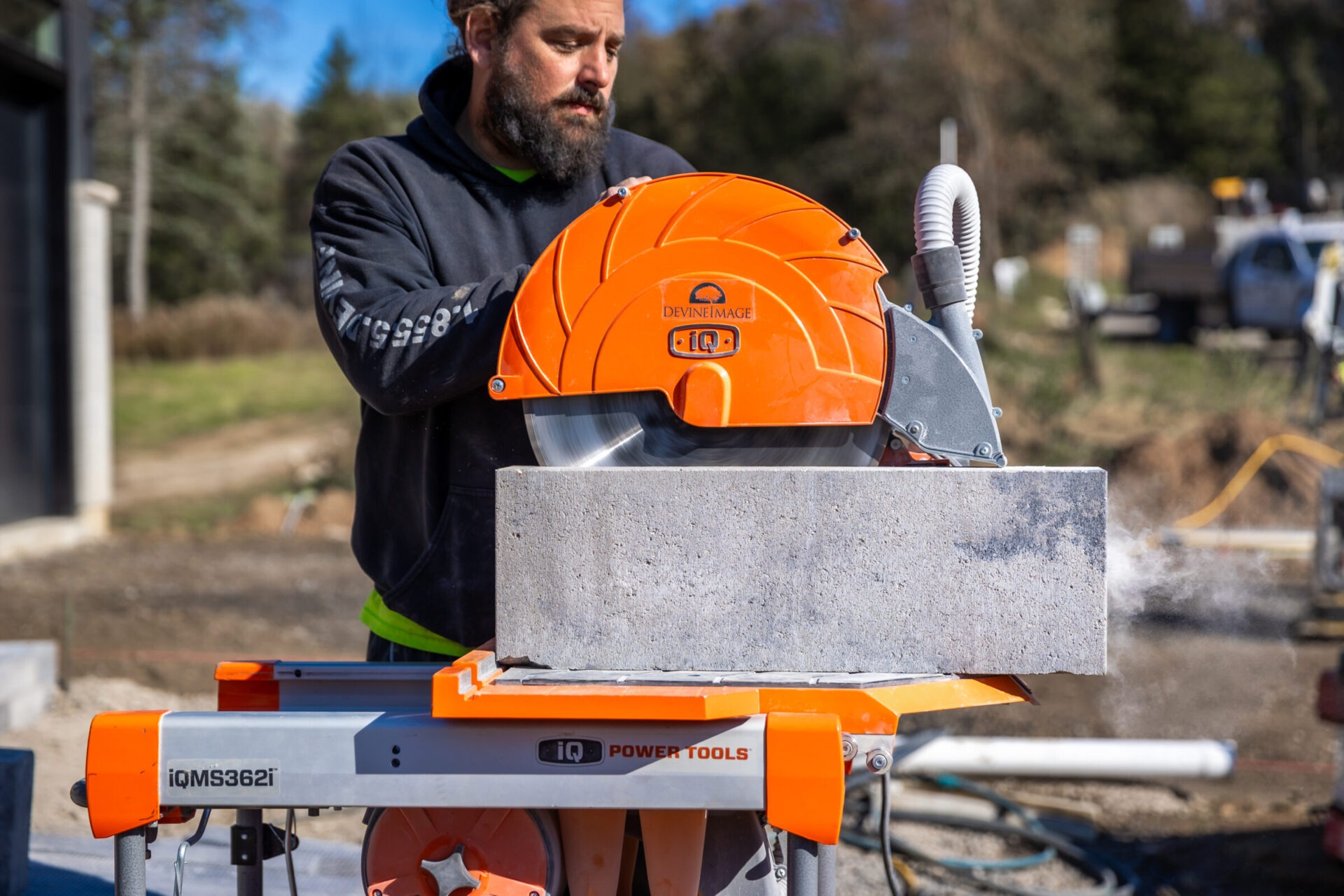 A person is cutting a cinder block with an orange masonry saw outdoors, with construction equipment and a vehicle in the background.