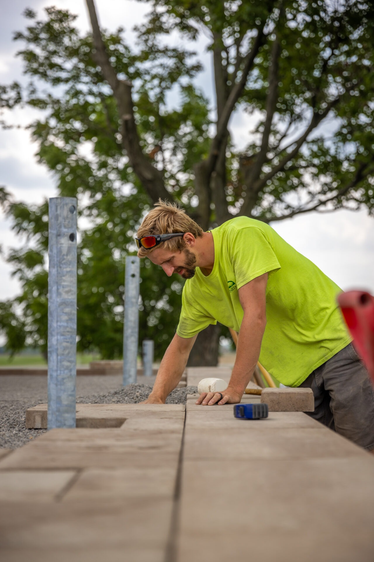 A person wearing a bright green shirt and safety glasses is laying paving stones. Outdoor setting with trees in the background. Construction environment.