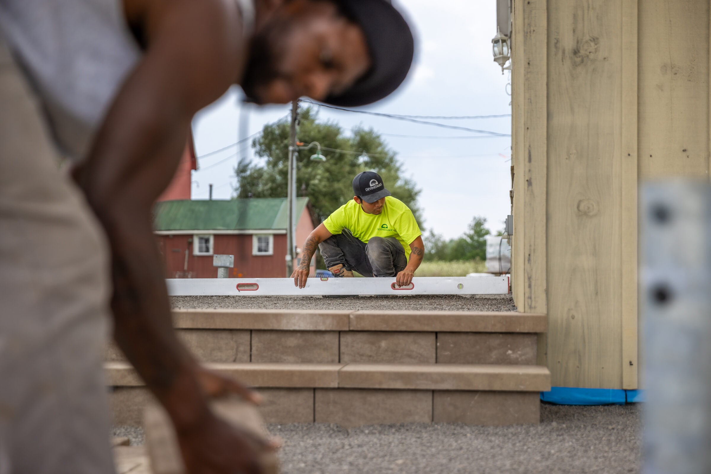 A person in a neon shirt and cap uses a level on steps outdoors, with another individual's silhouette partially visible in the foreground.