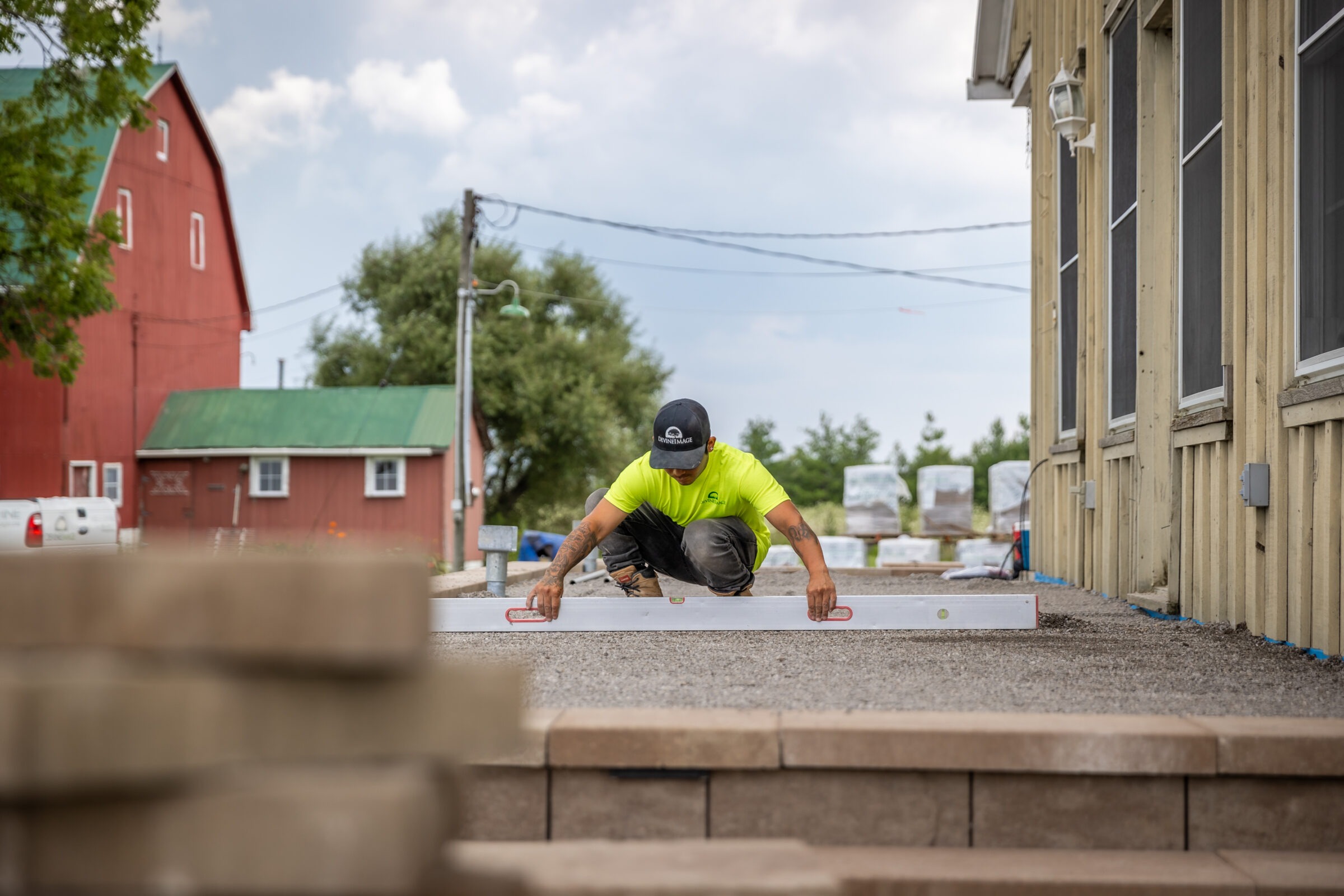 A person kneels on a concrete platform, measuring with a level, near a red barn, in a construction site, wearing high visibility clothing and a cap.