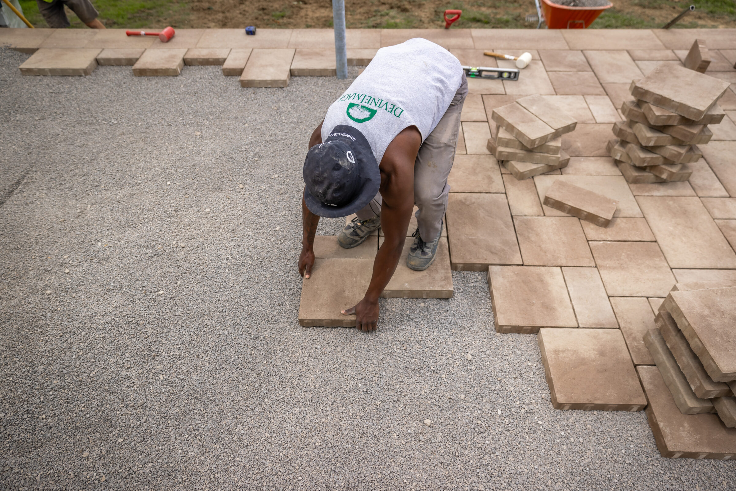A person is laying large paving stones to create a patio. Construction tools and materials are scattered around the work area.