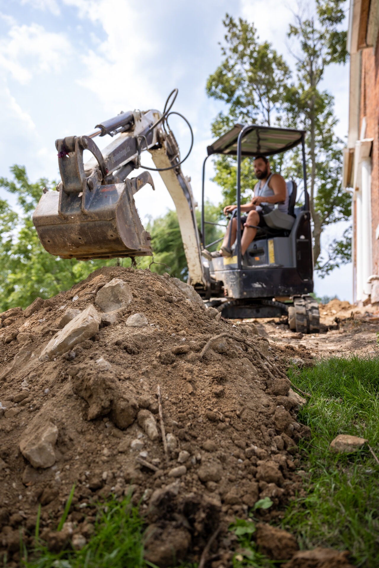 A person is operating an excavator near a mound of dirt on a construction site, with trees and a partially visible building in the background.