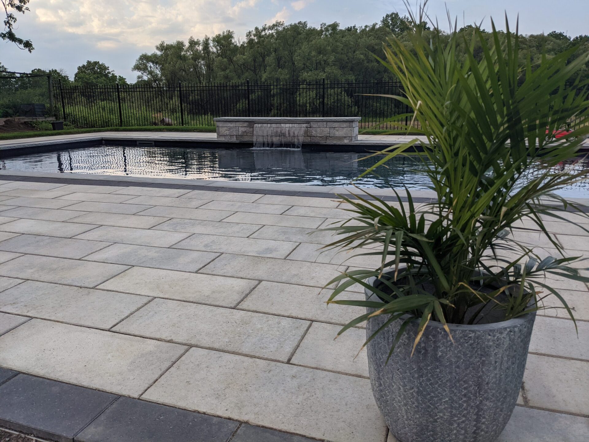 An outdoor swimming pool surrounded by a paved area and a metal fence. A potted plant is in the foreground with trees in the background.