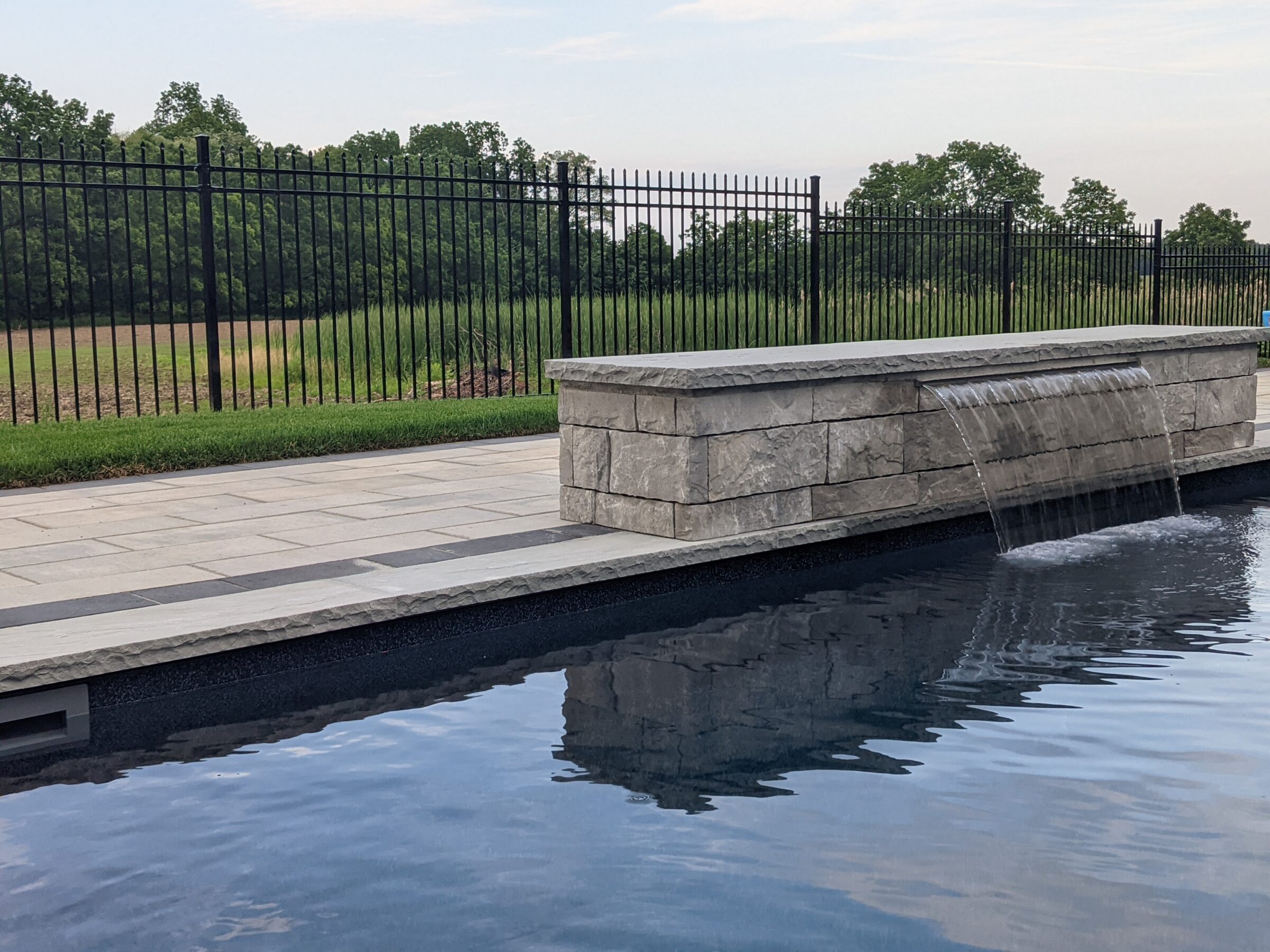This image shows an outdoor scene featuring a sleek rectangular pool with a stone water feature, neatly tiled patio, and a wrought-iron fence against a treelined horizon.