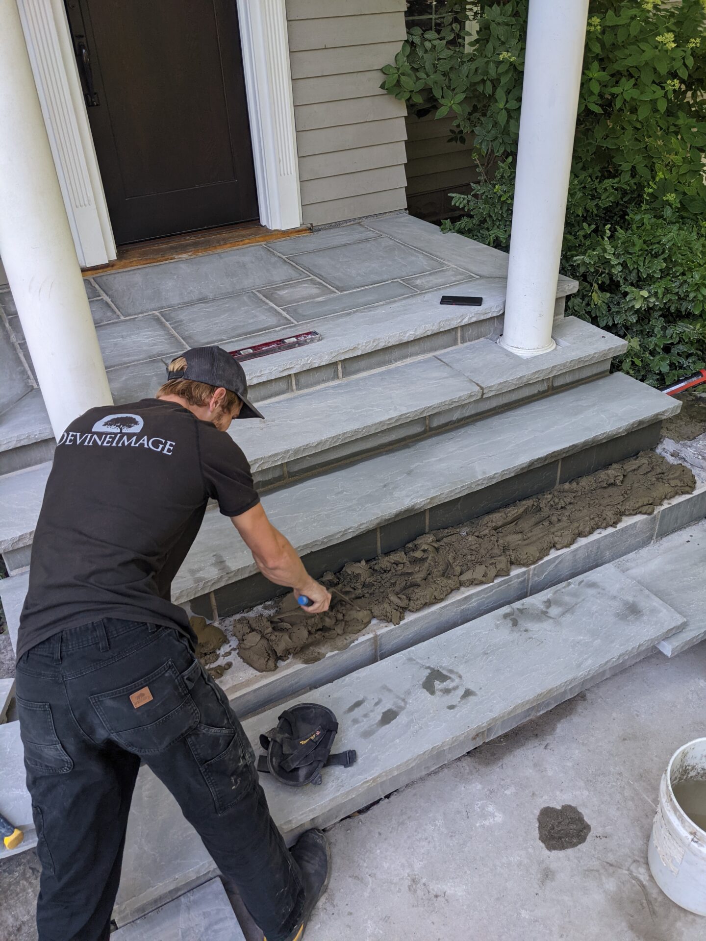 A person is applying mortar for tiles on outdoor steps near a white column and a black door, wearing a dark shirt and jeans, with tools nearby.