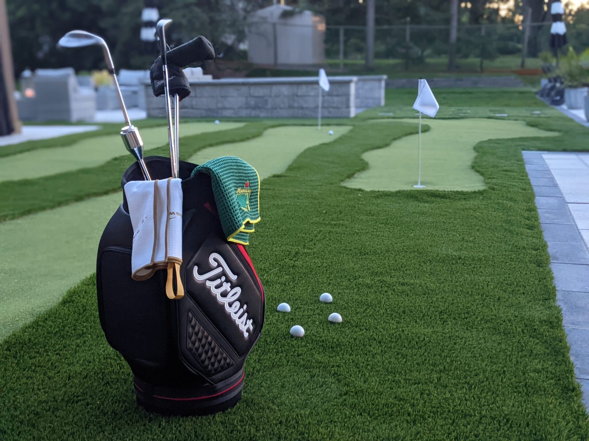 A golf bag with clubs and a white towel is positioned on artificial turf with golf balls nearby. Putting green with flagsticks in the background.