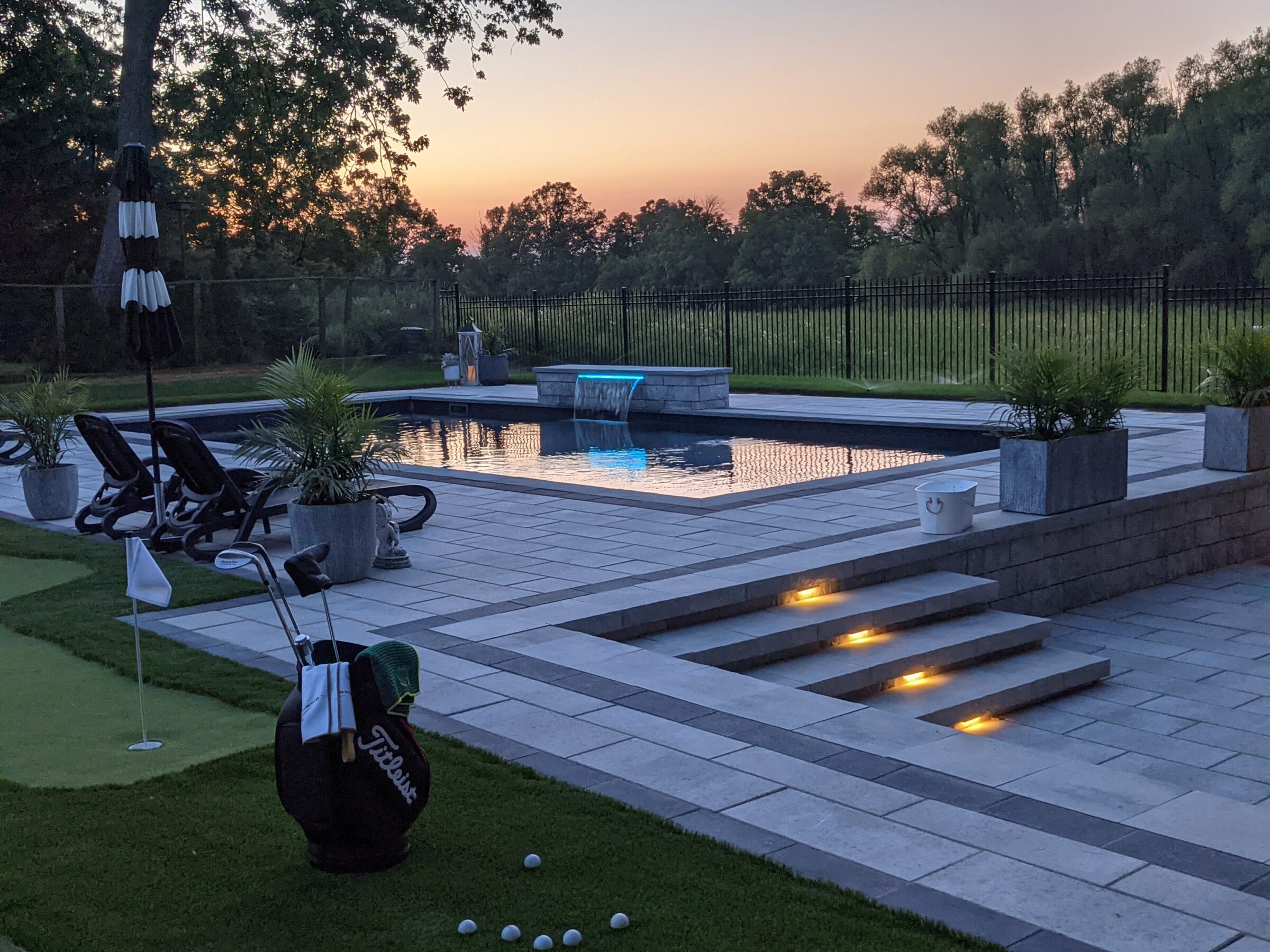 A tranquil backyard at dusk featuring a swimming pool, golf practice area, lounge chairs, lit steps, plants, and a view of trees against a sunset sky.