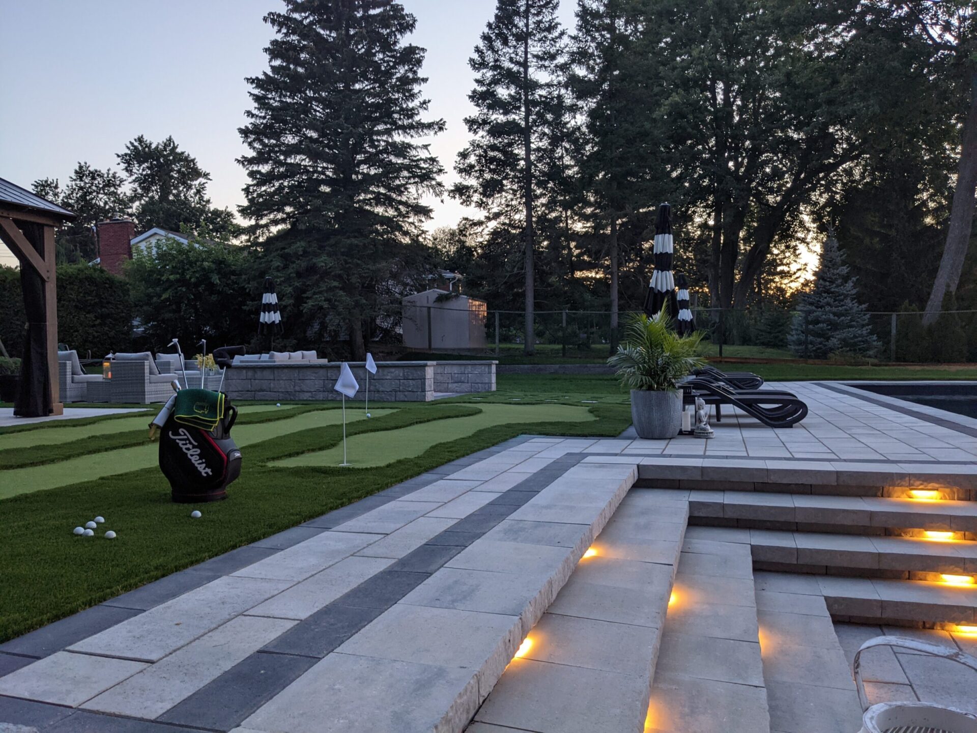 A luxurious backyard with a mini-golf setup, steps with integrated lights, a golf bag, loungers, and a tranquil early evening atmosphere.