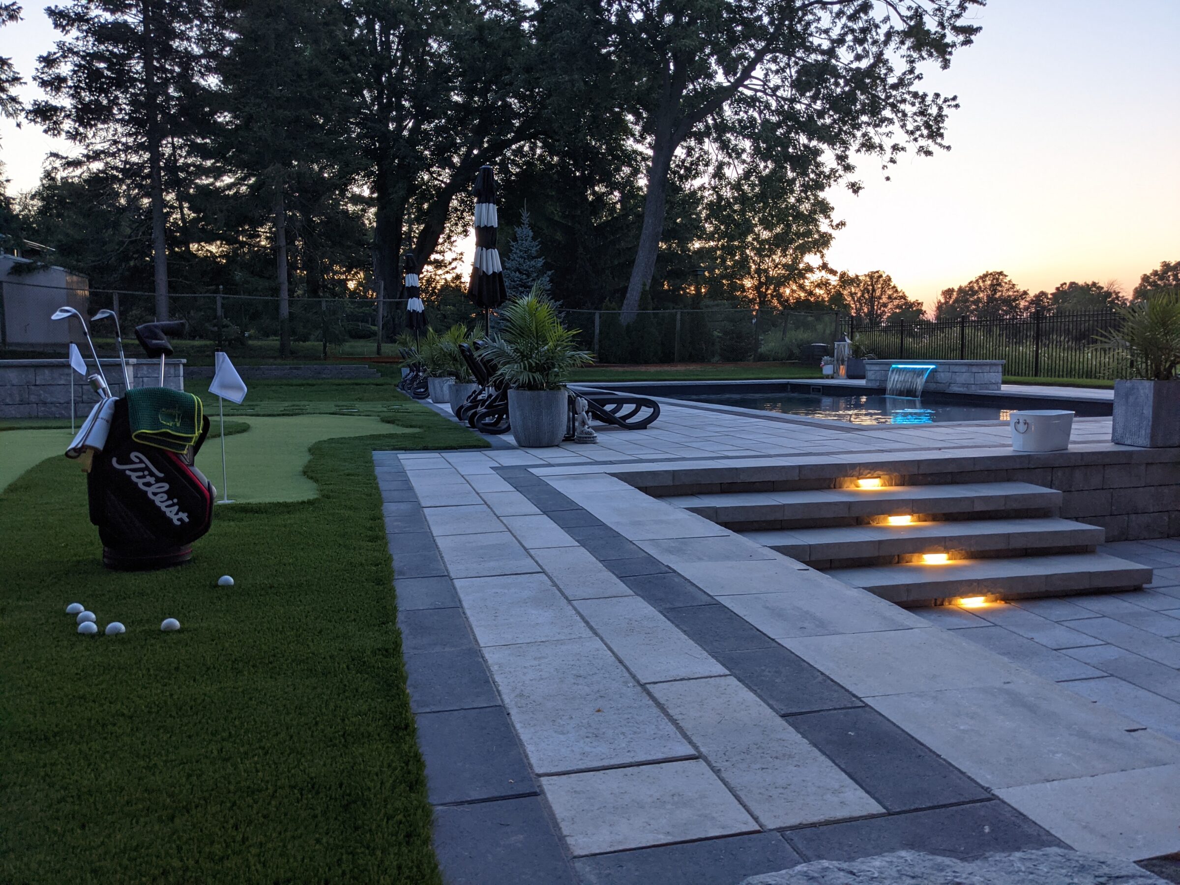 An artificial putting green with golf equipment near a swimming pool with a lit waterfall feature, stone steps with embedded lights, and outdoor furniture at dusk.