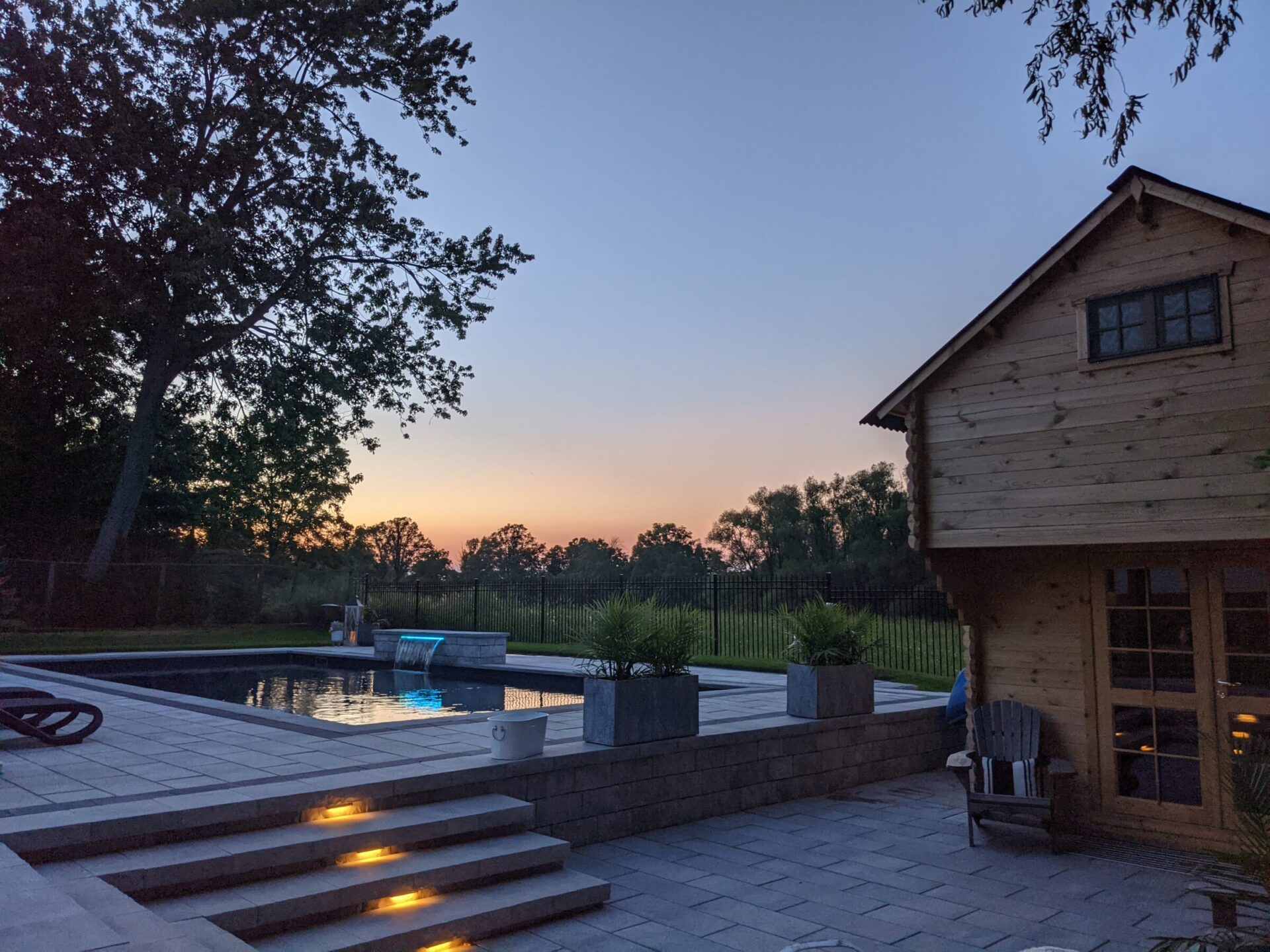 This image features a serene backyard at dusk with a wooden cabin, swimming pool, lit steps, a tree, and a colorful sky.