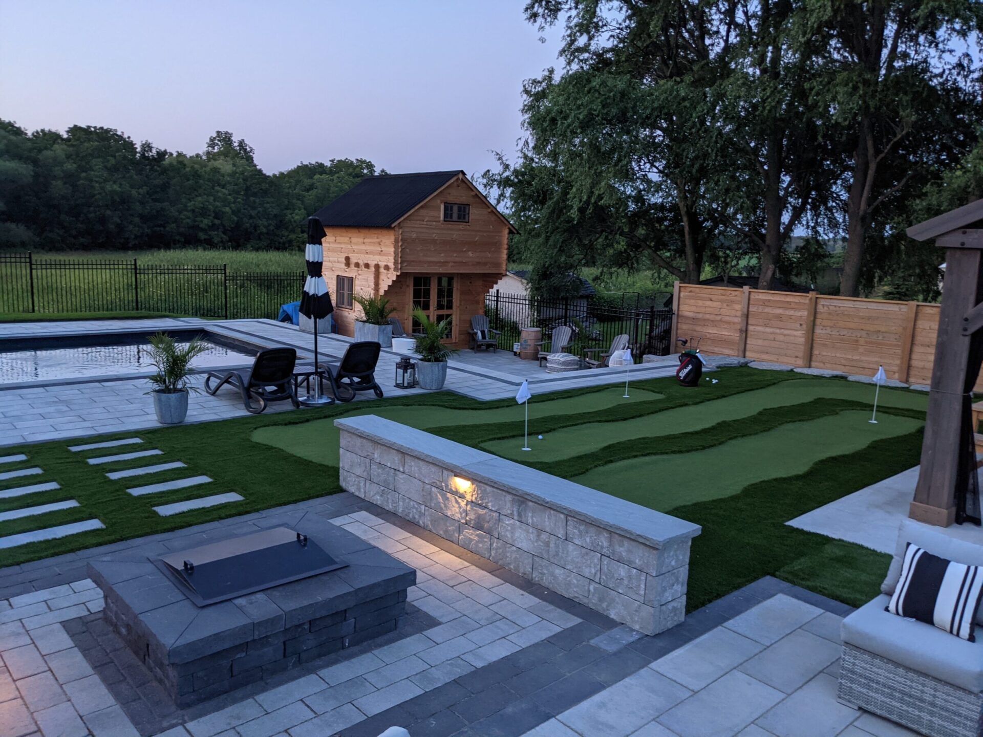 An elegant outdoor setting featuring a pool, patio with fire pit, lounging chairs, a putting green, and a wooden building at dusk.