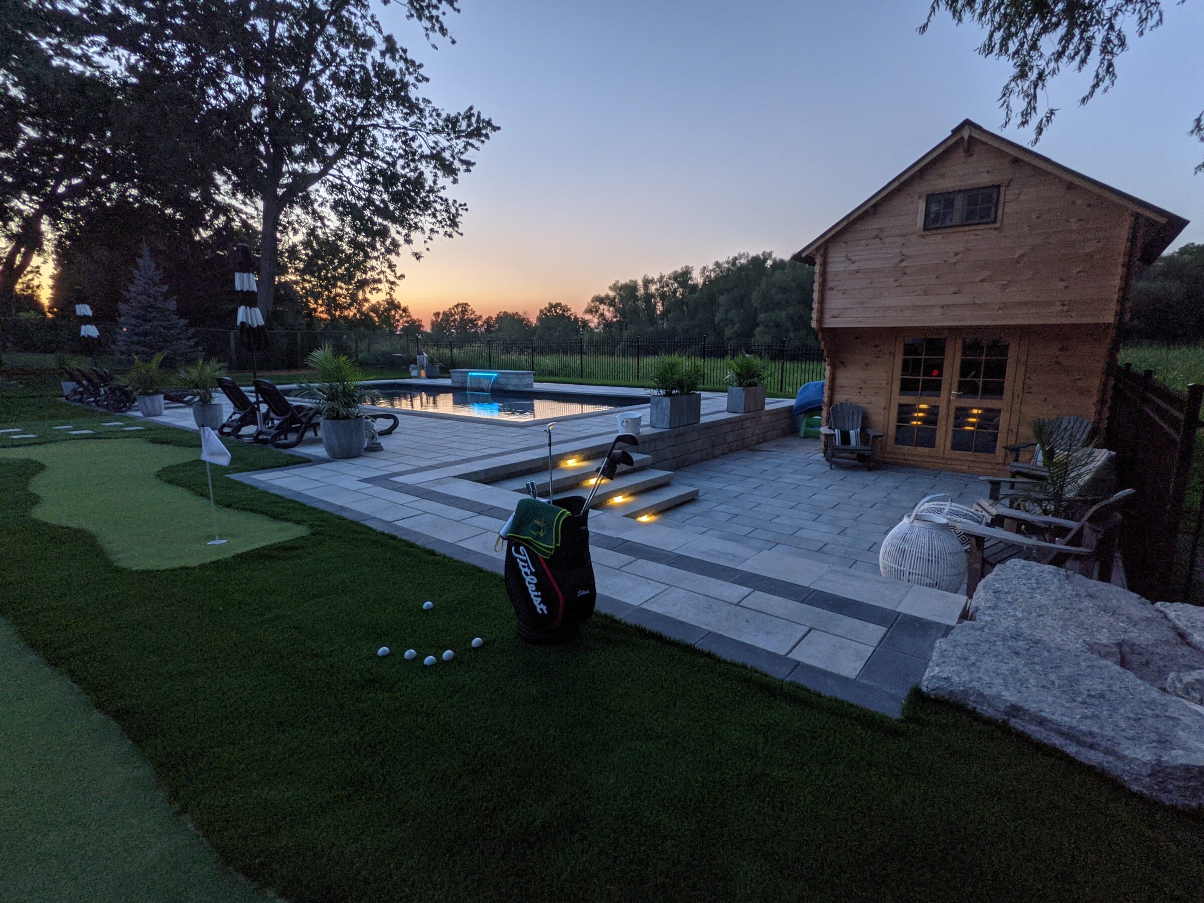 A luxurious backyard at dusk featuring a putting green, swimming pool, lounge chairs, and a cozy wooden cabin, all under a serene sky.
