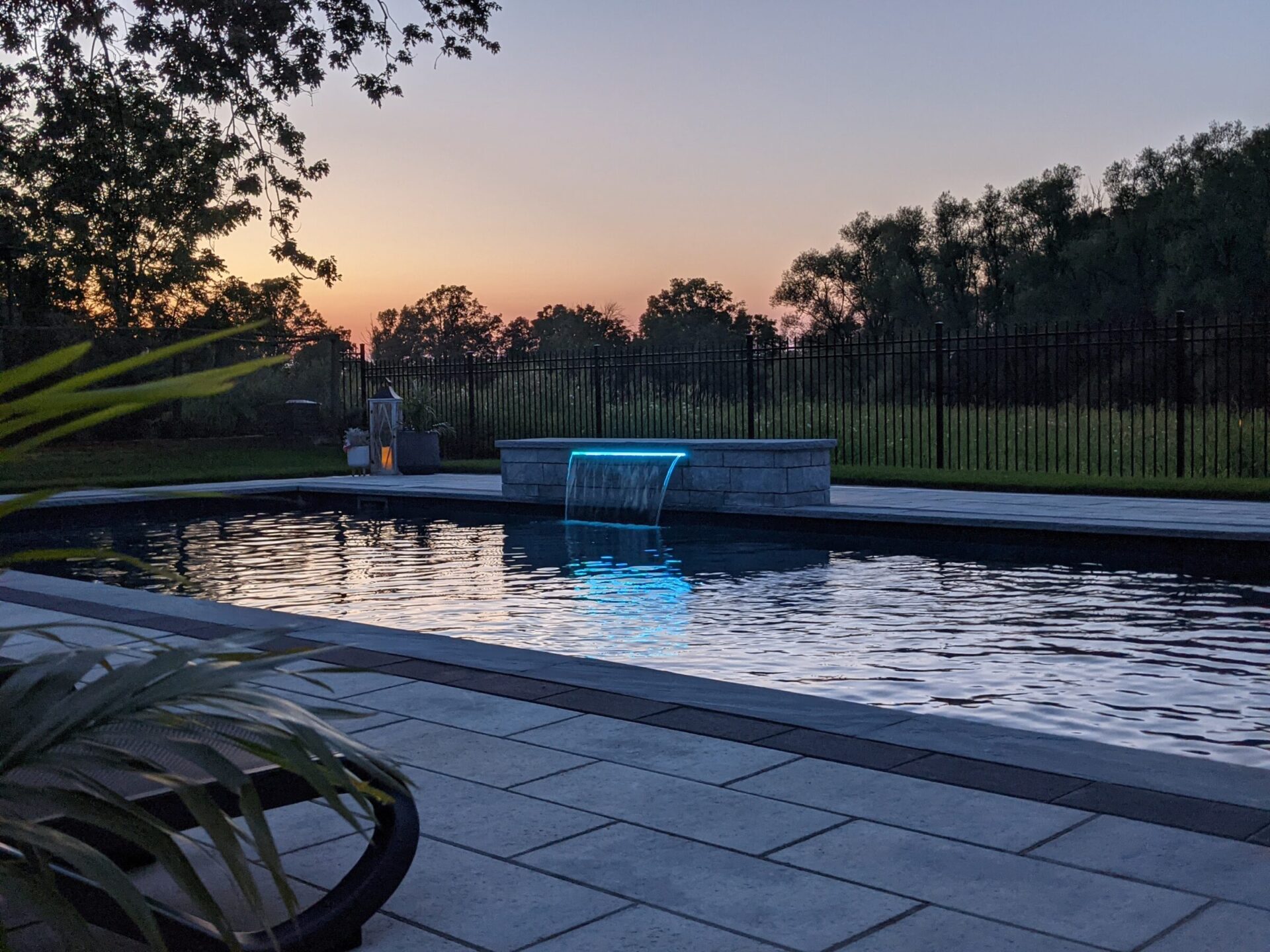 Evening by a serene backyard pool with blue illuminated water feature, overlooking a sunset sky beyond a wrought-iron fence lined with trees.