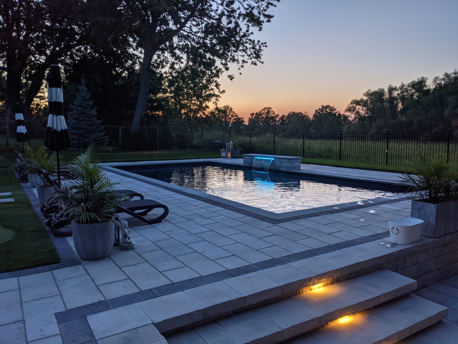 A serene backyard at dusk with a rectangular swimming pool, illuminated steps, lounging chairs, potted plants, and a sunset behind trees.