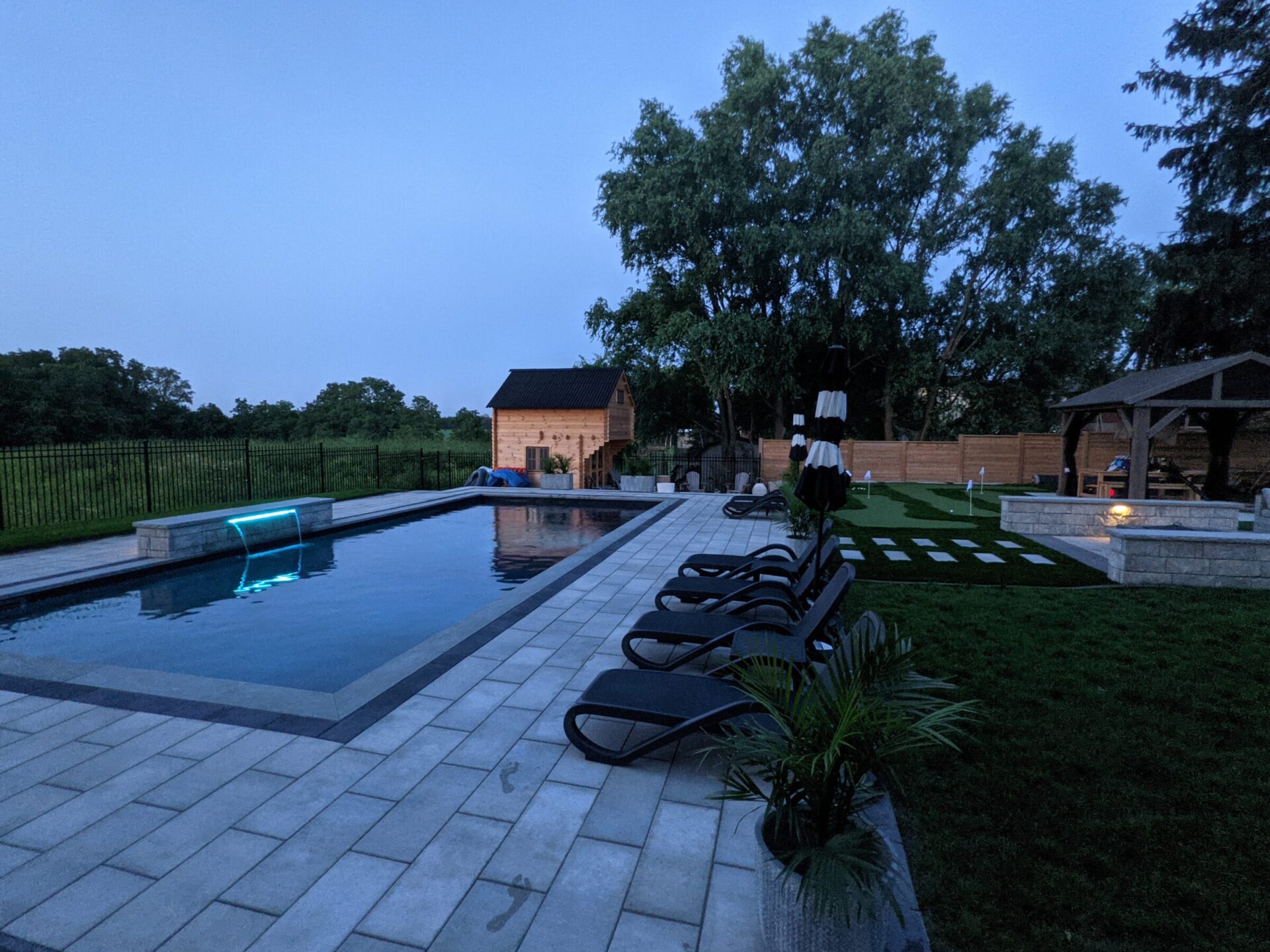 An outdoor pool with a neon blue light is featured at dusk. Lounge chairs, greenery, a gazebo, and a wooden fence complement the tranquil setting.