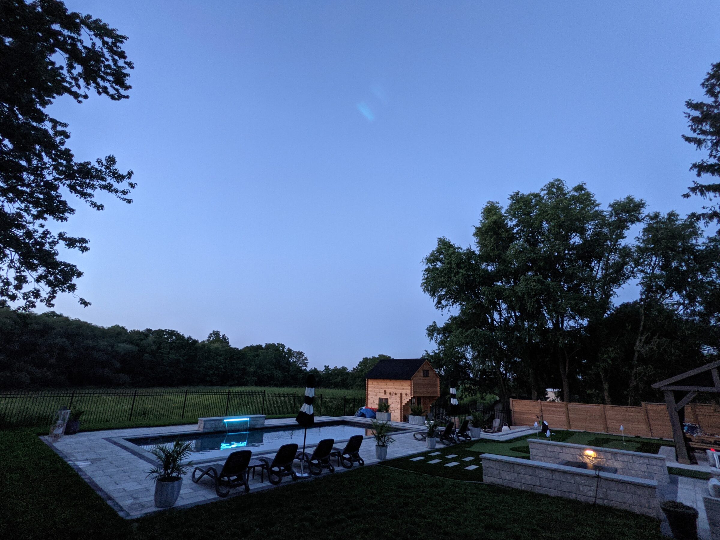 A serene twilight scene featuring an illuminated pool, a well-manicured lawn, outdoor furniture, and trees, with a soft gradient sky overhead.