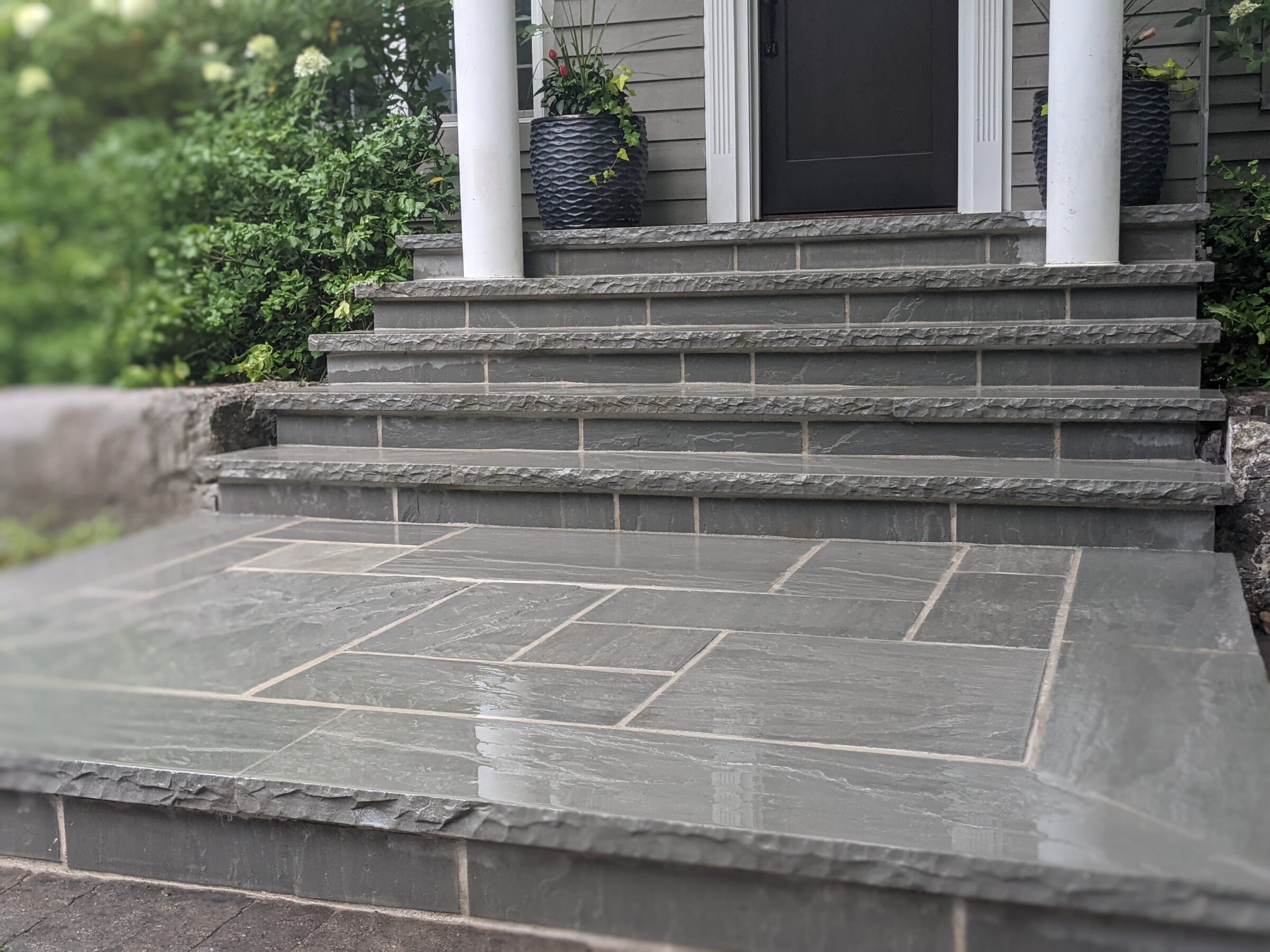 A home entrance featuring stone steps leading to a dark door, flanked by white columns and potted plants, with green foliage in the background.