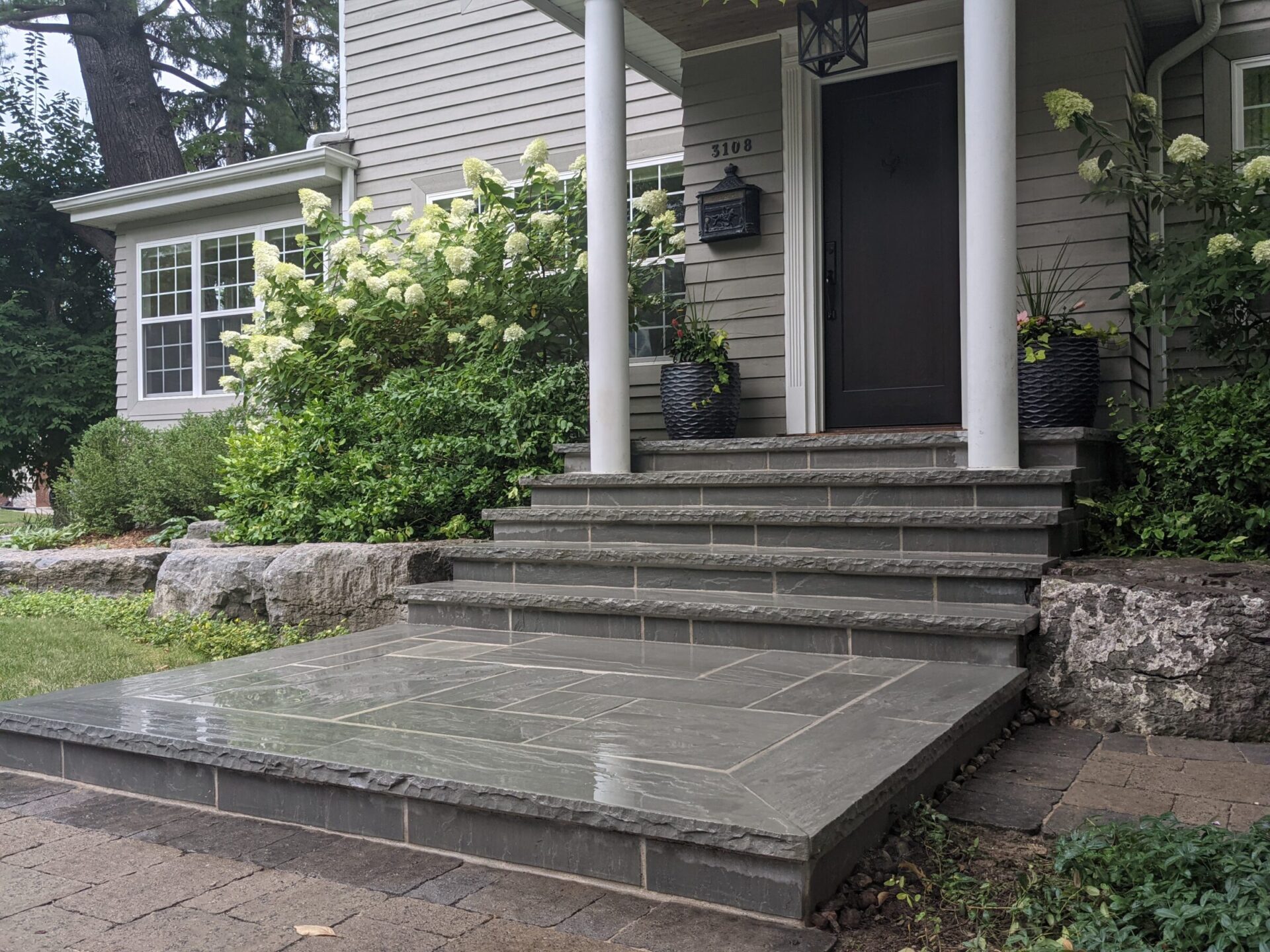 The image shows the front porch of a house with stairs, a dark door, white columns, hydrangeas, and potted plants, presenting a welcoming, tidy entrance.