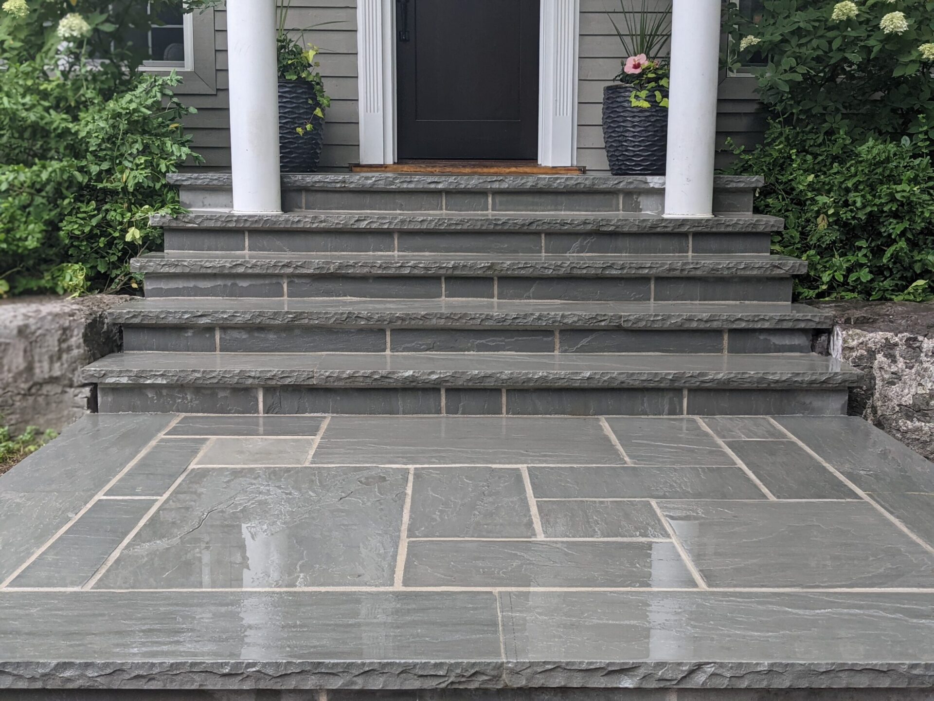 The image shows a stone tiled entryway leading to a set of stairs and a door flanked by columns, with potted plants and greenery beside the entrance.