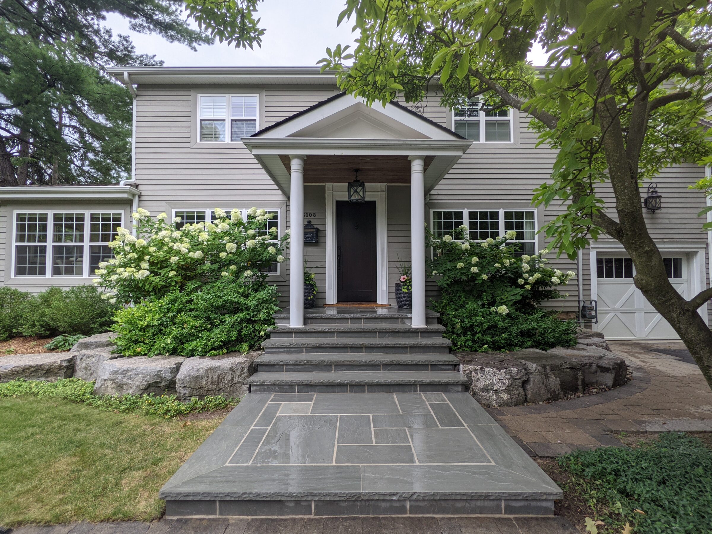 A grey two-story house with white trim, a covered entryway, blooming hydrangeas, stone steps, and a garage door, nestled among green trees.