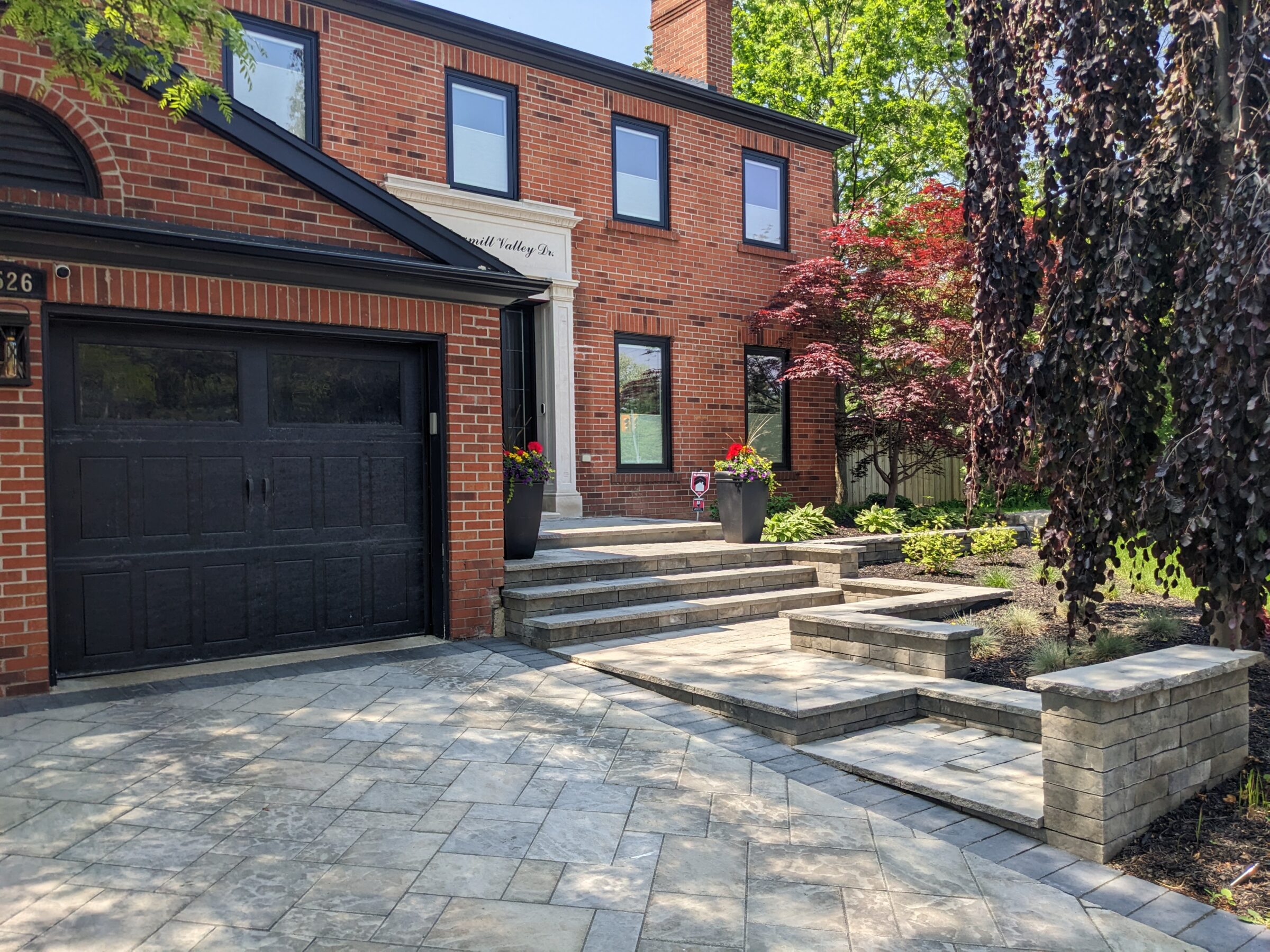 This image shows a two-story red brick house with black garage doors, a stone paver driveway, landscaped garden, and steps leading to the front door.