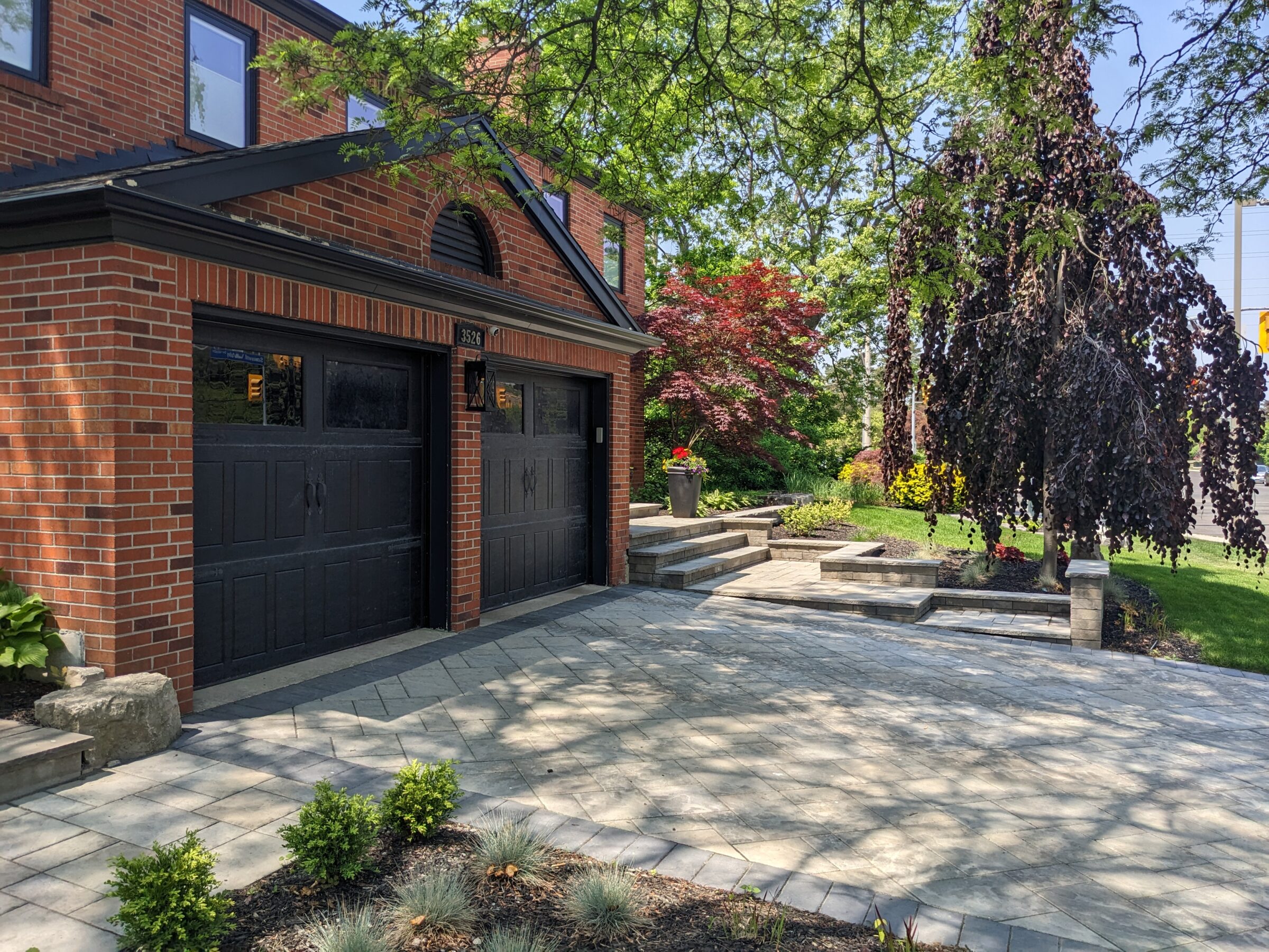 This image shows a two-car garage with black doors on a brick residence. A landscaped garden with a stone pathway and lush trees under a blue sky.