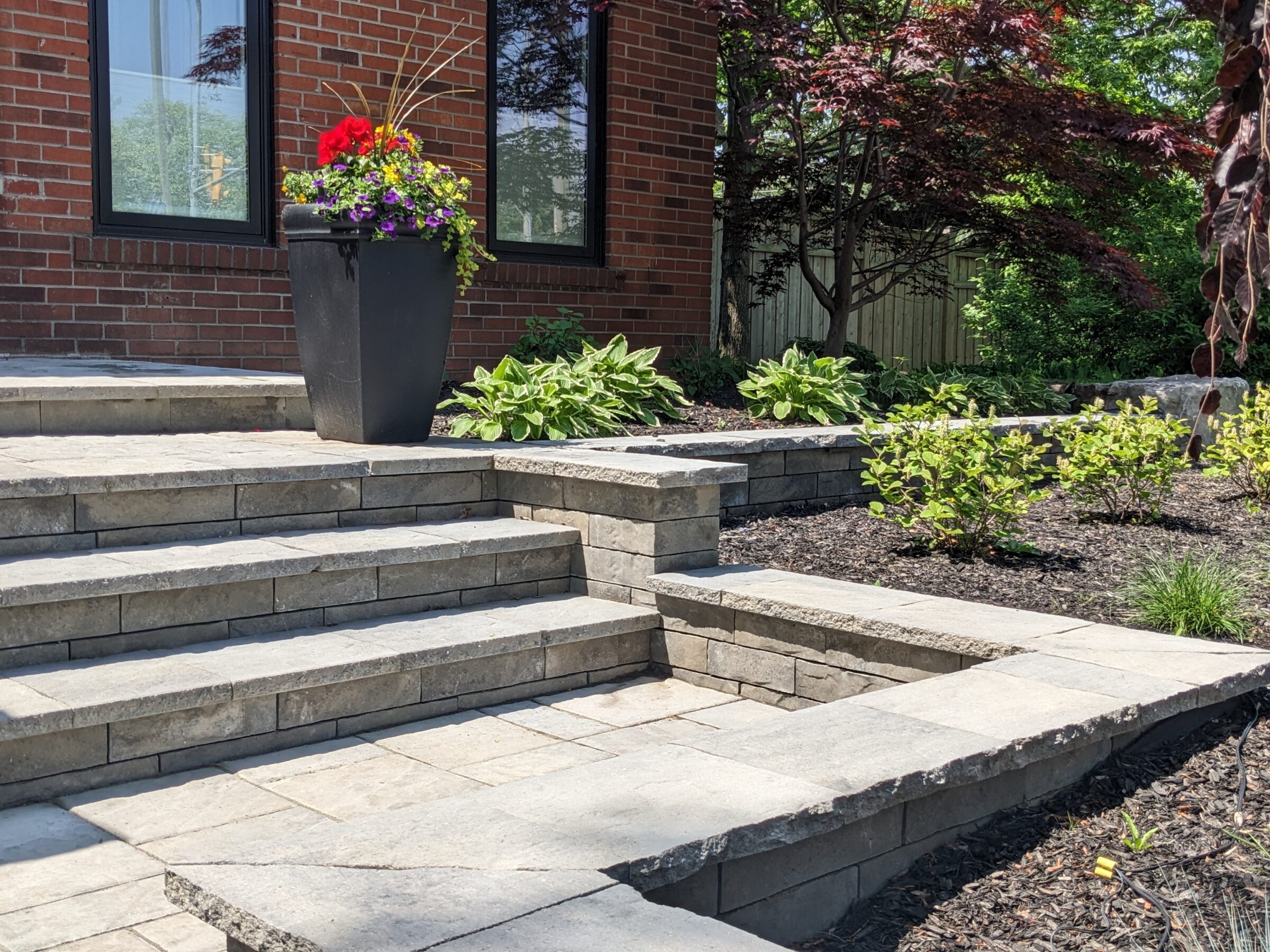 A landscaped yard featuring stone steps, a large container with colorful flowers, various plants, and a red-brick building with reflective windows in the background.