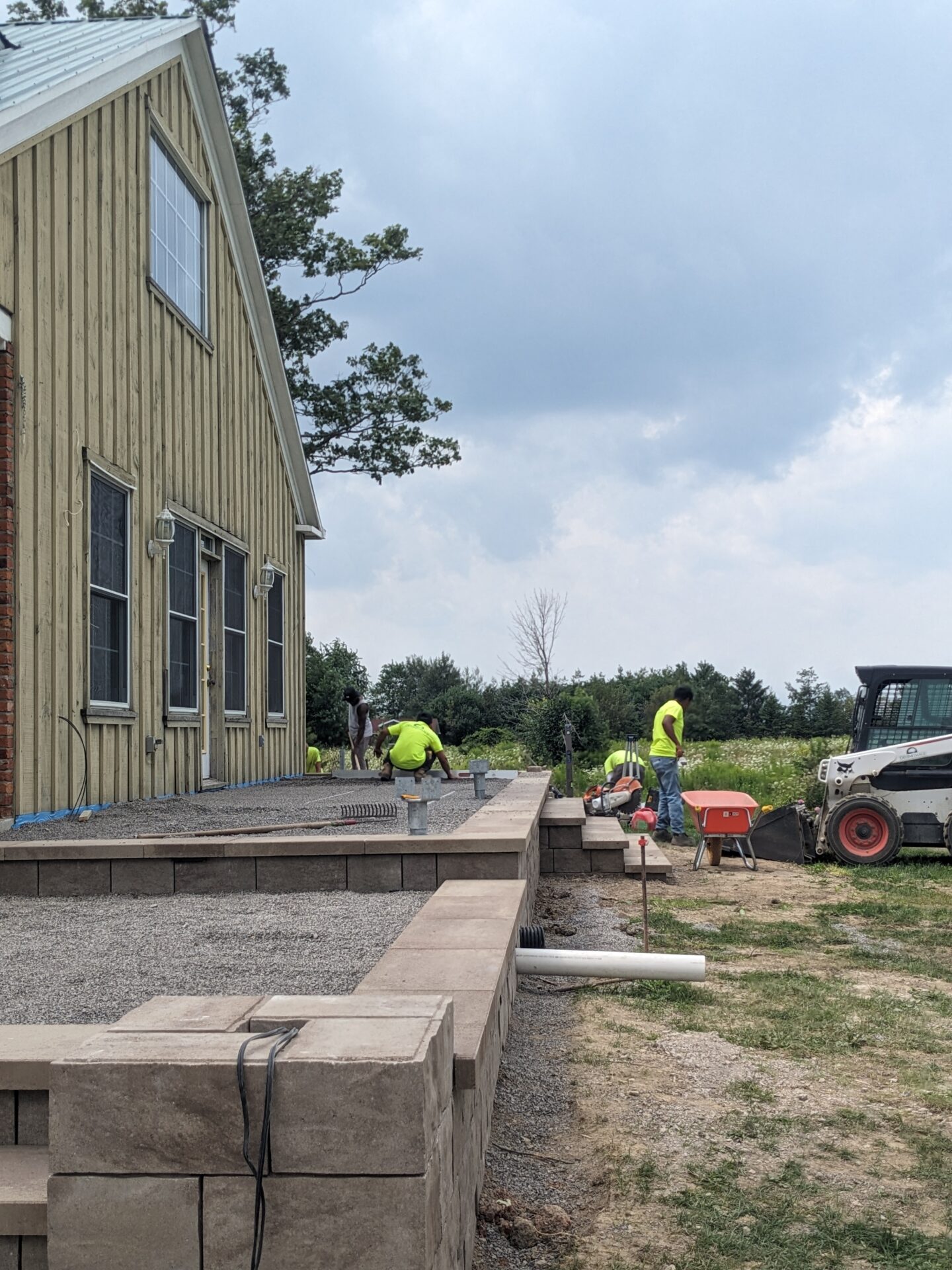 Several people in high-visibility shirts are working on a construction site near a wooden building, handling materials on a gravel base under a cloudy sky.